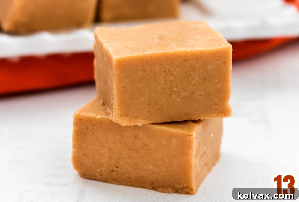 A close-up shot of two perfectly stacked pieces of creamy Easy Peanut Butter Fudge, resting on a clean white surface with a platter full of more fudge in the soft-focused background.