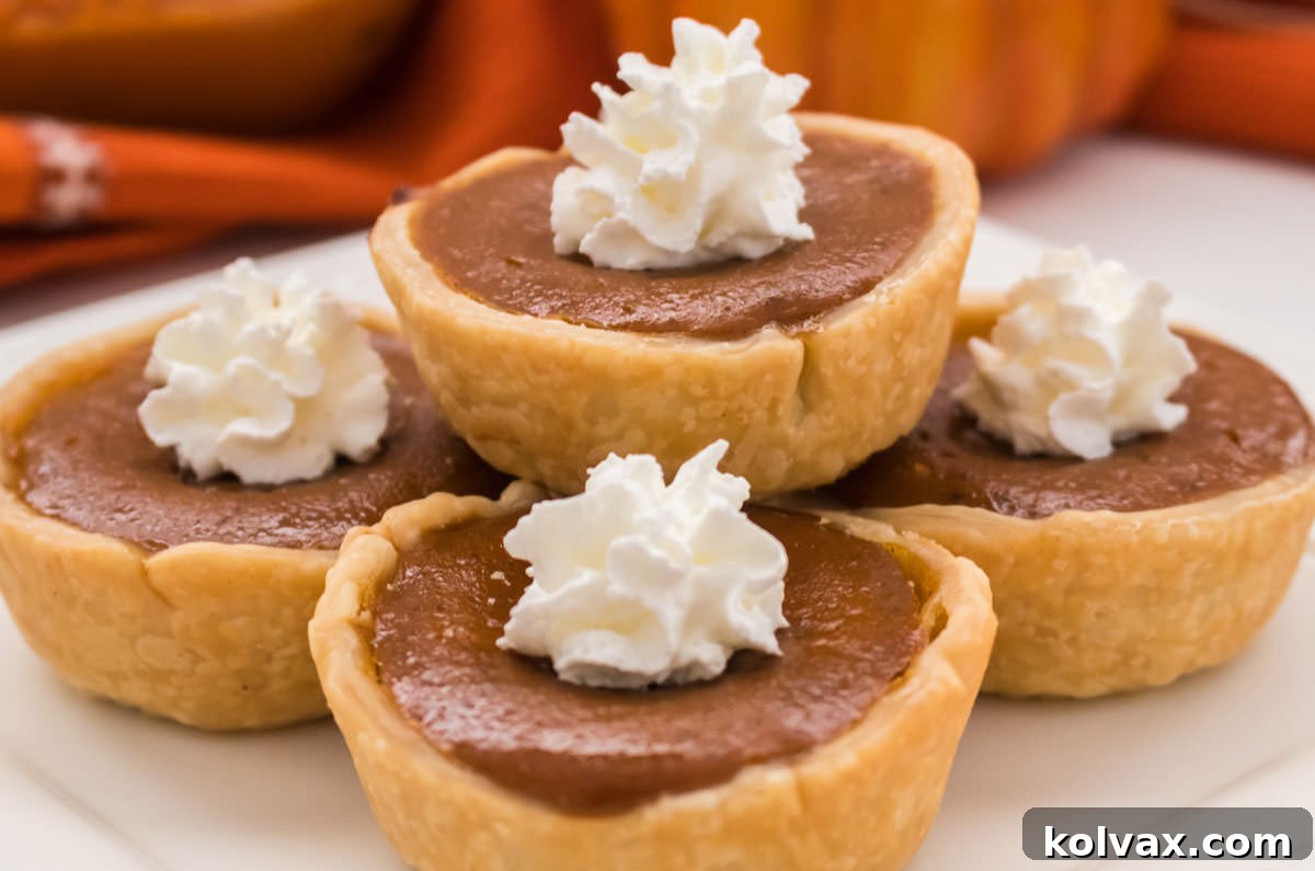 Closeup on a stack of five Mini Pumpkin Pies, topped with dollops of whipped cream, sitting on a white plate. Each pie is perfectly golden with a rich filling.