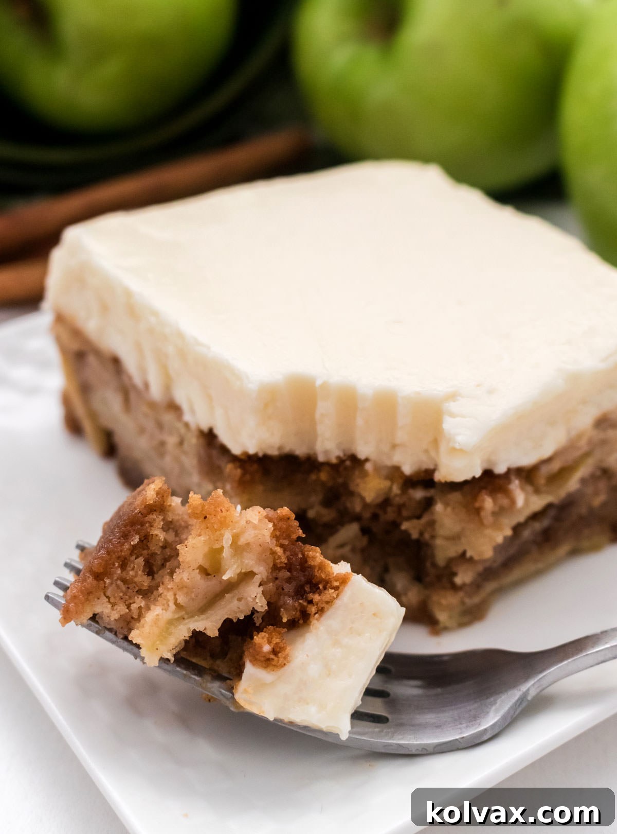 Orchard Apple Cake with Cream Cheese Crown 11 A close-up shot of a single slice of Apple Cake, generously frosted with cream cheese, resting on a white plate. A fork with a perfect bite of cake is positioned enticingly in front of the slice, highlighting the moist cake and creamy frosting.