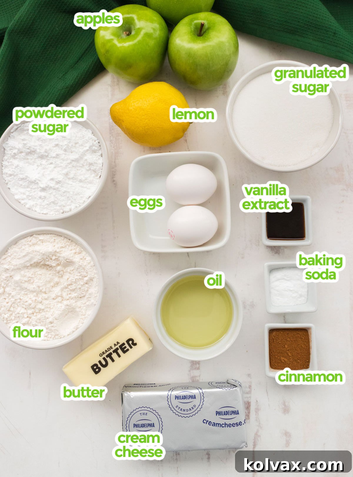 Orchard Apple Cake with Cream Cheese Crown 3 An overhead shot showcasing all the essential ingredients laid out for making Apple Cake. Visible items include fresh green Granny Smith apples, a bright yellow lemon, granulated sugar, powdered sugar, eggs, a bottle of vanilla extract, baking soda, ground cinnamon, cooking oil, all-purpose flour, sticks of butter, and a block of cream cheese – everything needed for a complete, homemade apple cake and its frosting.