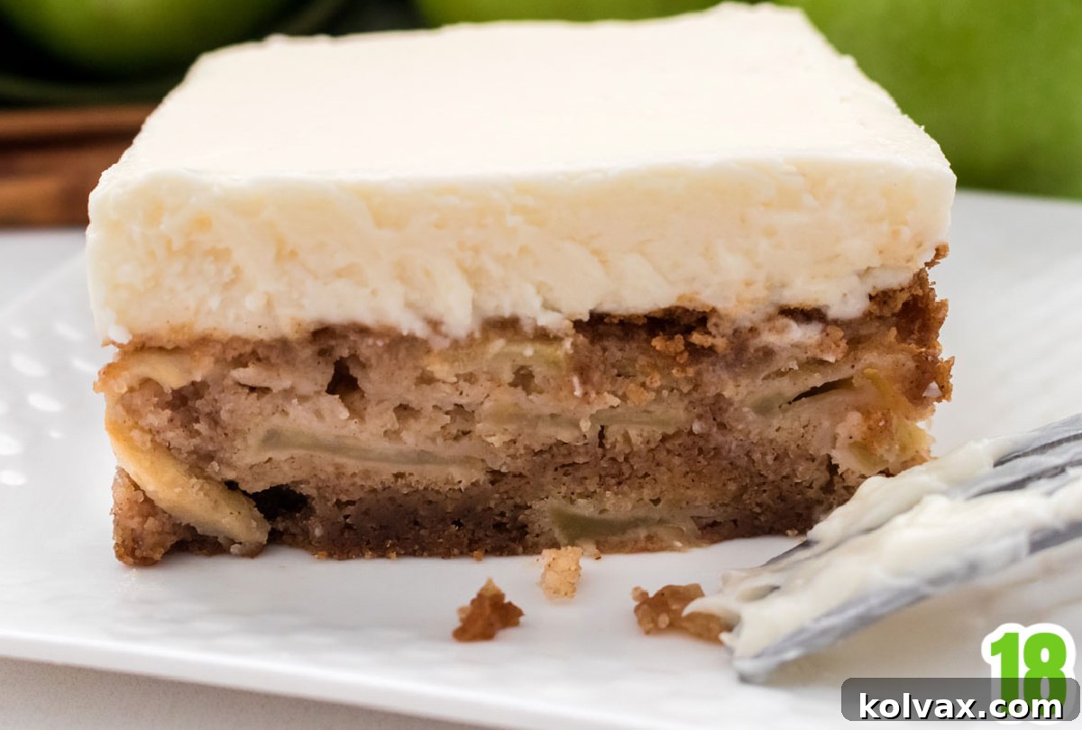 Orchard Apple Cake with Cream Cheese Crown 10 A close-up, side-view photograph of a slice of Apple Cake, clearly showcasing the distinct layers of tender apple pieces embedded within the moist cake, topped with a generous, thick layer of creamy white cream cheese frosting. The focus is on the beautiful texture and composition of the cake.