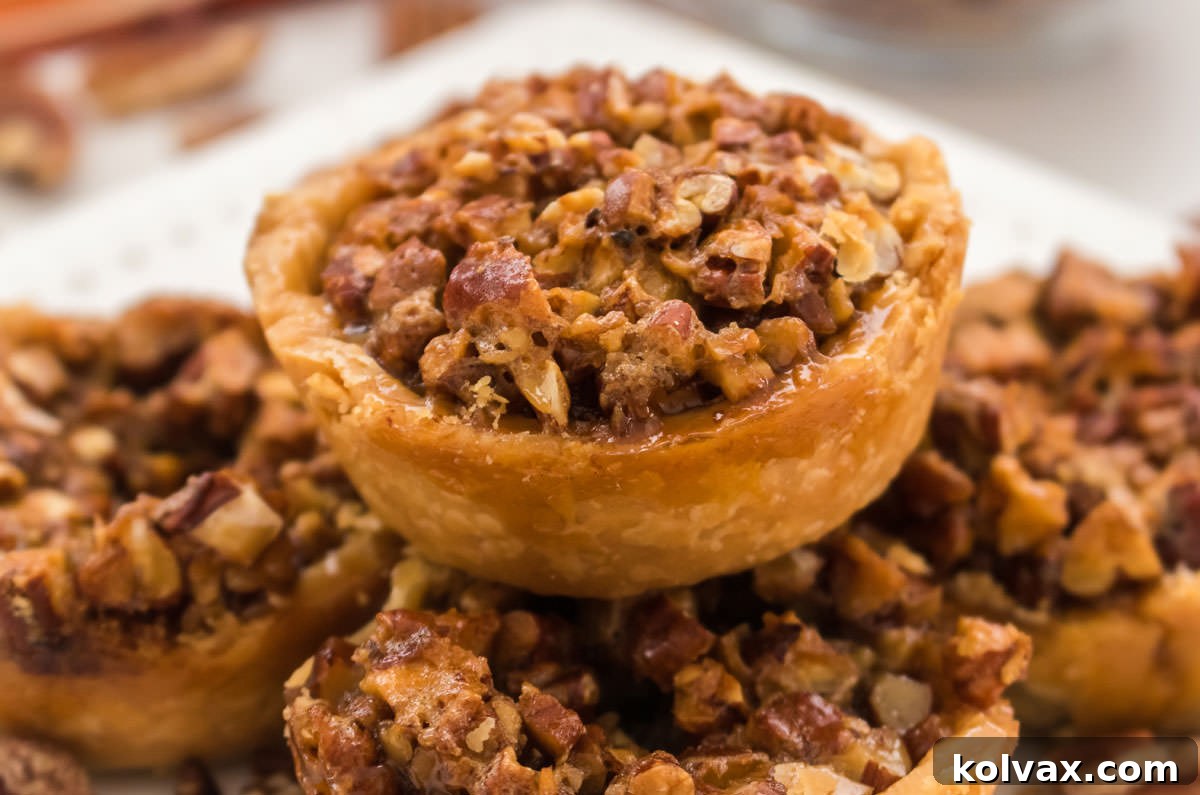 Closeup on a Mini Pecan Pie sitting atop a stack of mini pies on a white plate, showcasing its golden crust and glistening pecan topping.