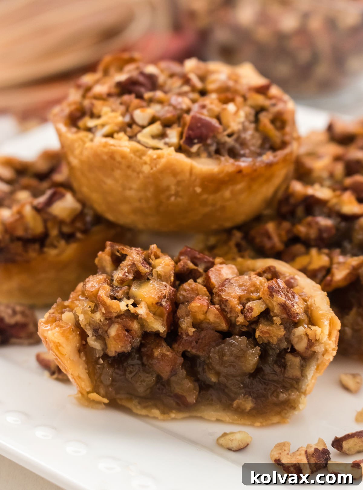 Closeup on five perfectly baked Mini Pecan Pies sitting on a white plate in front of a glass bowl filled with chopped pecans, ready to be enjoyed.