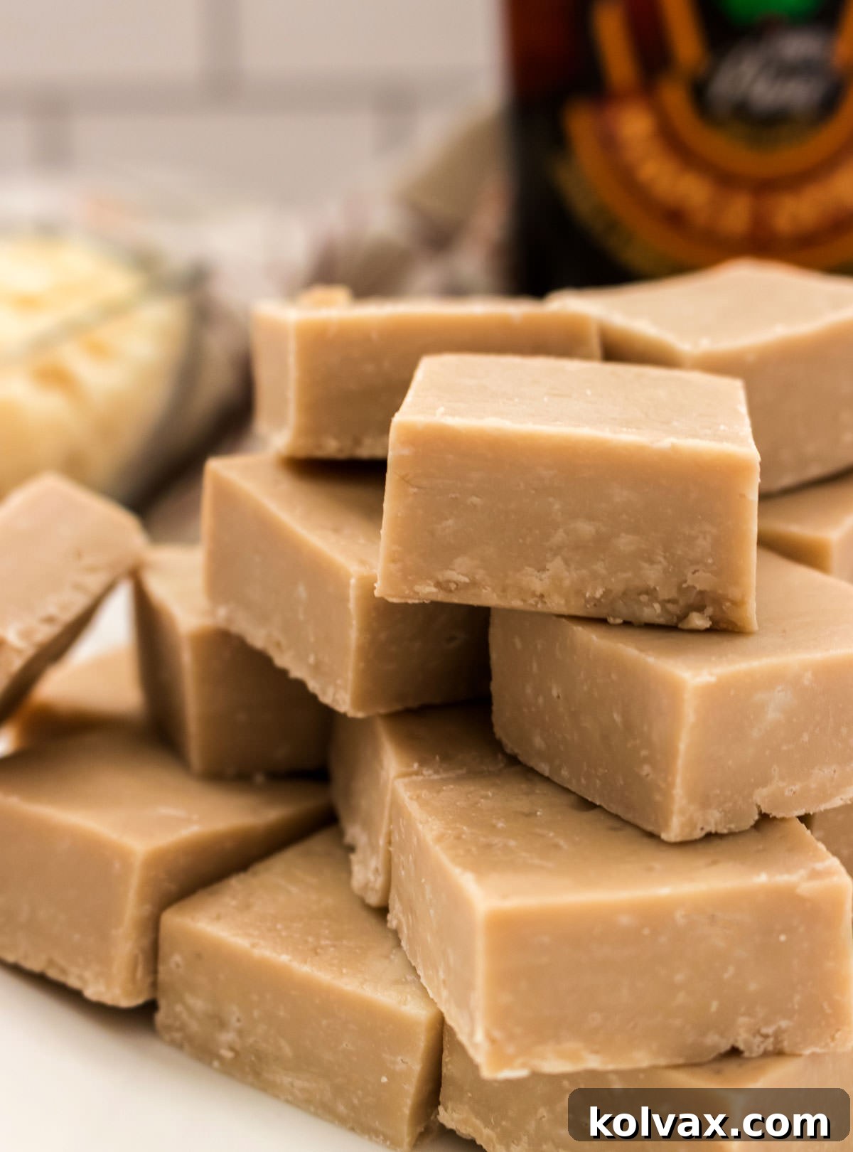 Closeup on a stack of Maple Fudge sitting on a white surface in front of a bottle of maple syrup and a glass bowl filled with White Chocolate Chips.