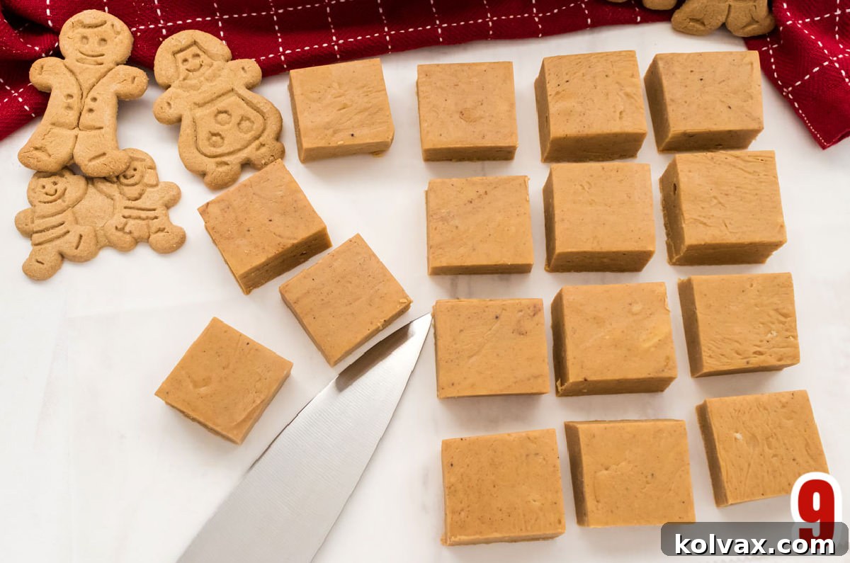 Sixteen neatly cut pieces of Gingerbread Fudge arranged in rows, alongside festive gingerbread cookies and a silver knife, ready for serving on a white surface.