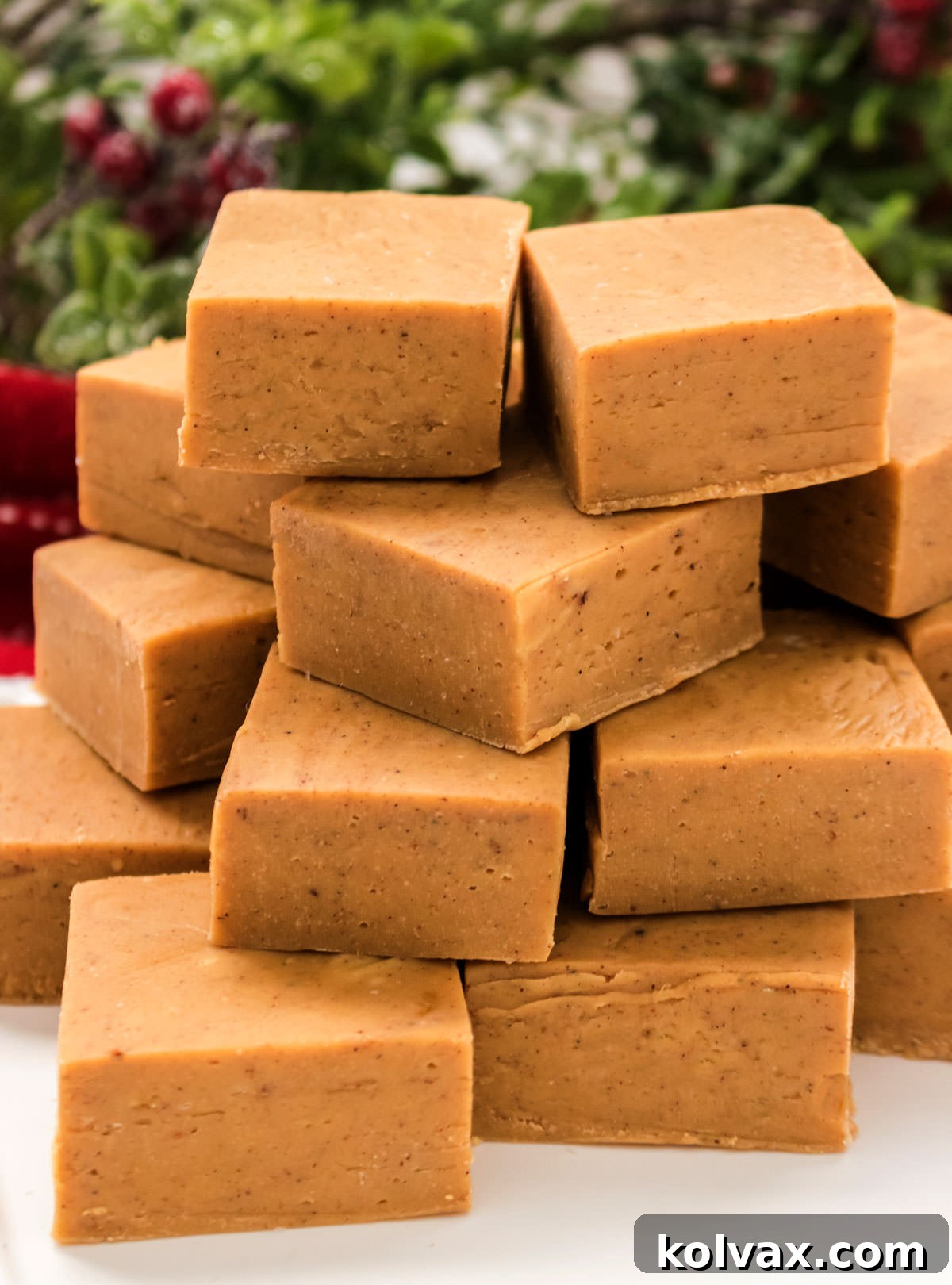 A close-up shot of a stack of Gingerbread Fudge pieces, glistening and inviting, set on a white table with elegant holiday greenery and holly leaves in the background, perfect for a festive display.