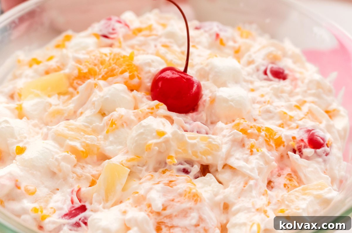 Closeup of a glass serving bowl filled with vibrant Ambrosia Salad, garnished with a single maraschino cherry with its stem, inviting guests to indulge in this sweet, creamy fruit concoction.