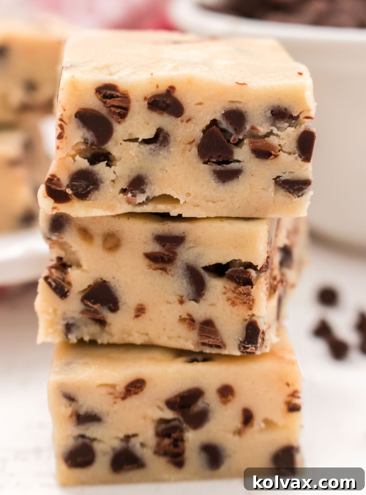 A close-up of three stacked Cookie Dough Fudge pieces, with more fudge on a plate in the background, all on a white surface.