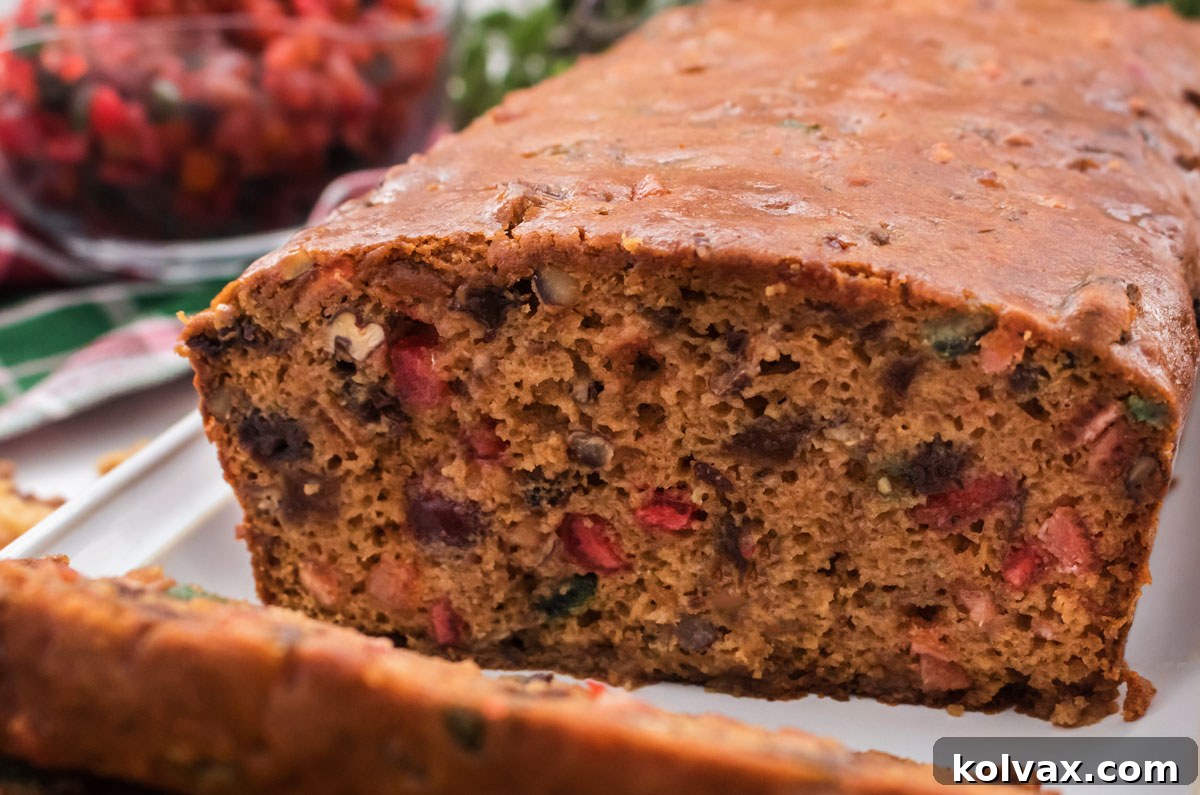 Closeup on a loaf of Fruit Cake sitting on a white platter with Christmas Decorations in the background. This homemade fruit cake is moist, delicious, and dense with rich fruity flavors, far superior to store-bought versions. Perfect for the holidays!