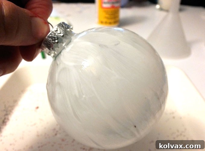 Sparkling Silver Christmas Tree Baubles 5 A craft brush applying Mod Podge evenly onto a clear glass Christmas tree ornament, preparing it for glitter application.