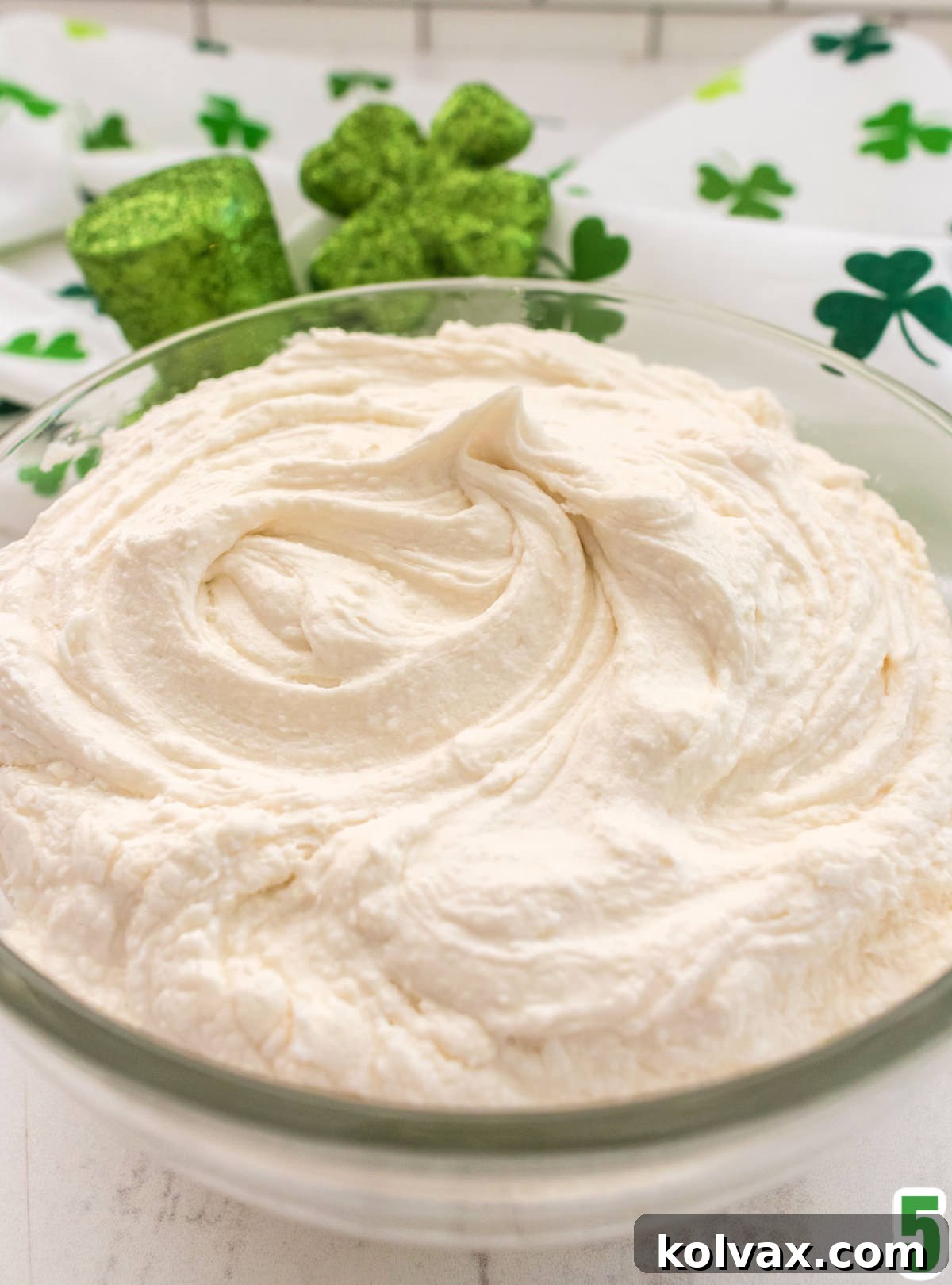 A pristine clear glass bowl generously filled with light, fluffy Whipped Vanilla Buttercream Frosting, showcasing its smooth and inviting texture.