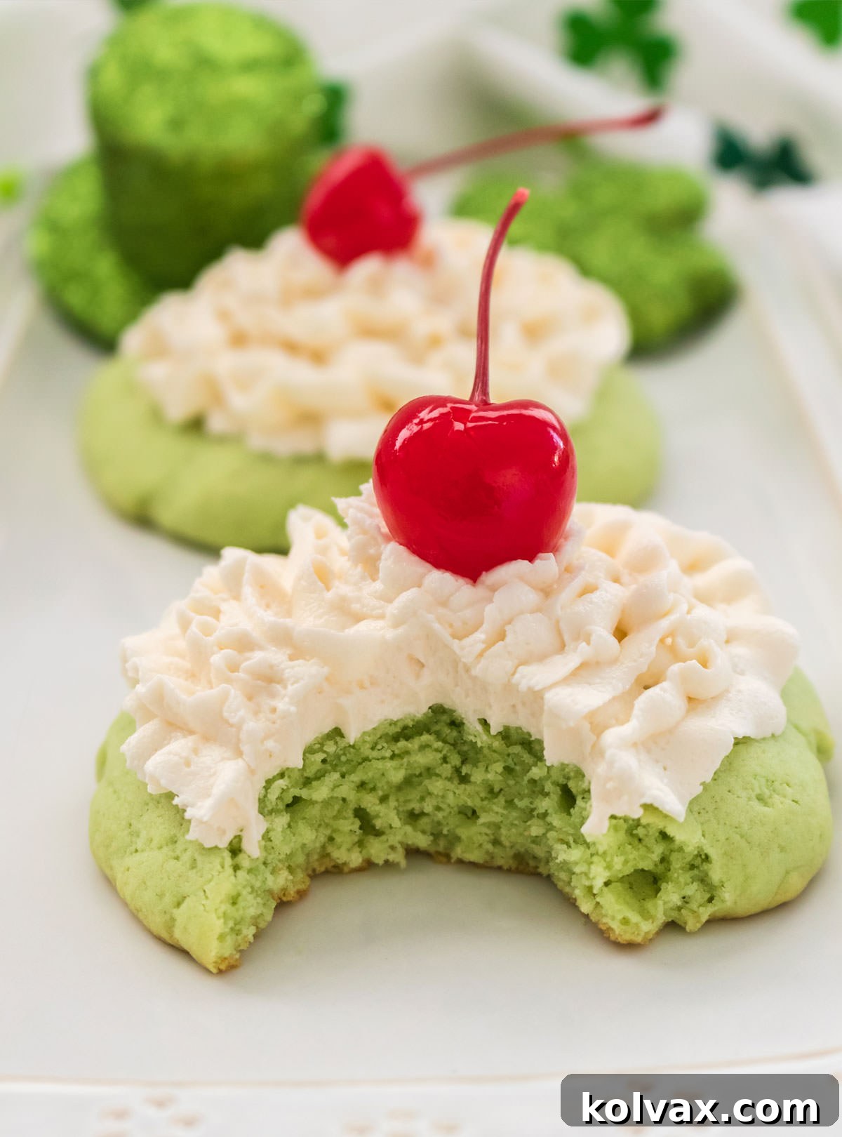 A charming white dessert platter showcasing two exquisite Shamrock Shake Cookies, one with an inviting bite taken out of it, revealing its fluffy interior.