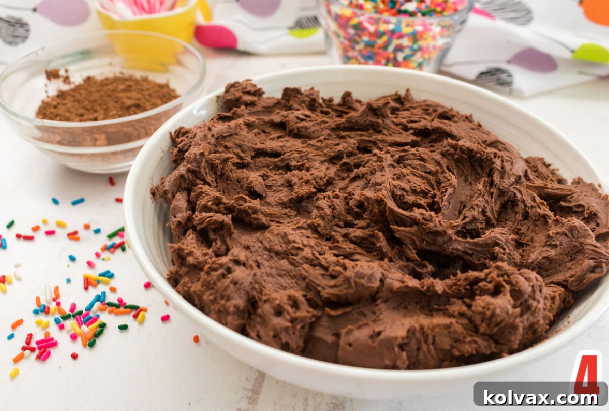 A pristine white bowl filled with glossy homemade Chocolate Buttercream Frosting, surrounded by sprinkles and a ramekin of cocoa powder, ready for decorating.