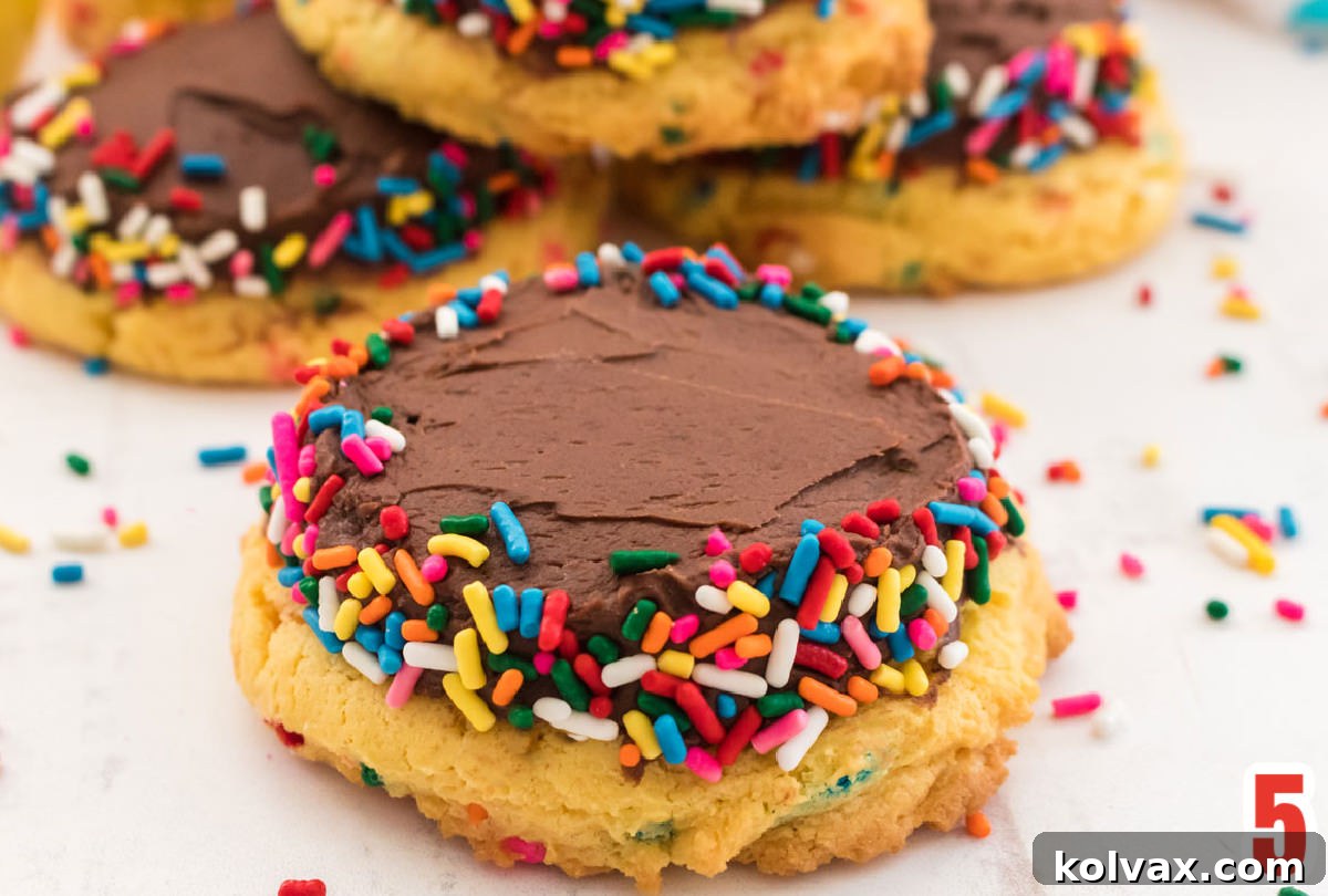 A delightful close-up of a frosted Birthday Cake Mix Cookie, adorned with sprinkles, resting on a white table surrounded by more cookies and festive decorations.