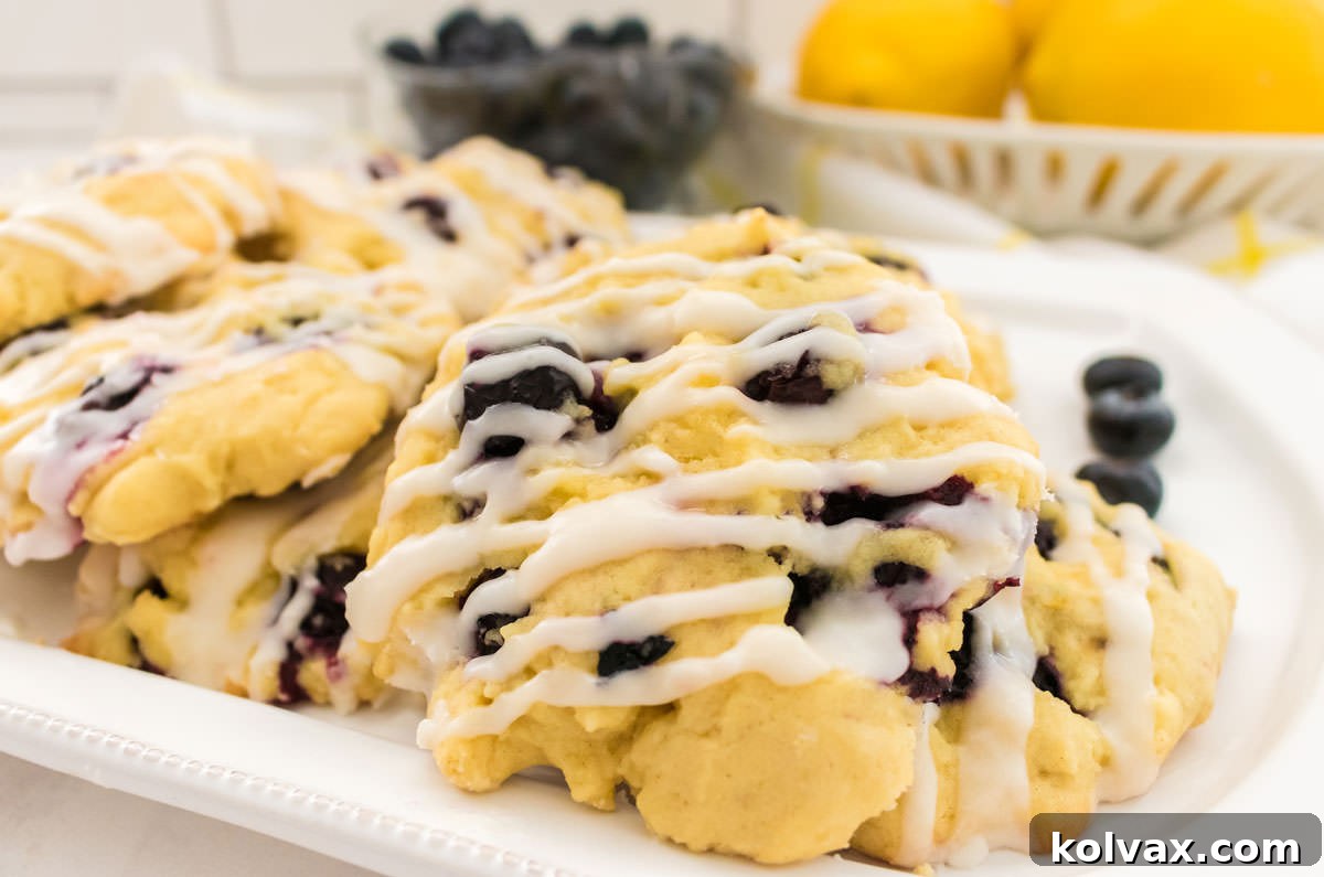 Closeup on a white platter filled with Lemon Blueberry Cookies sitting on a white table in front of a bowl of fresh blueberries and a bowl of fresh lemons. Each cookie is beautifully drizzled with lemon icing.