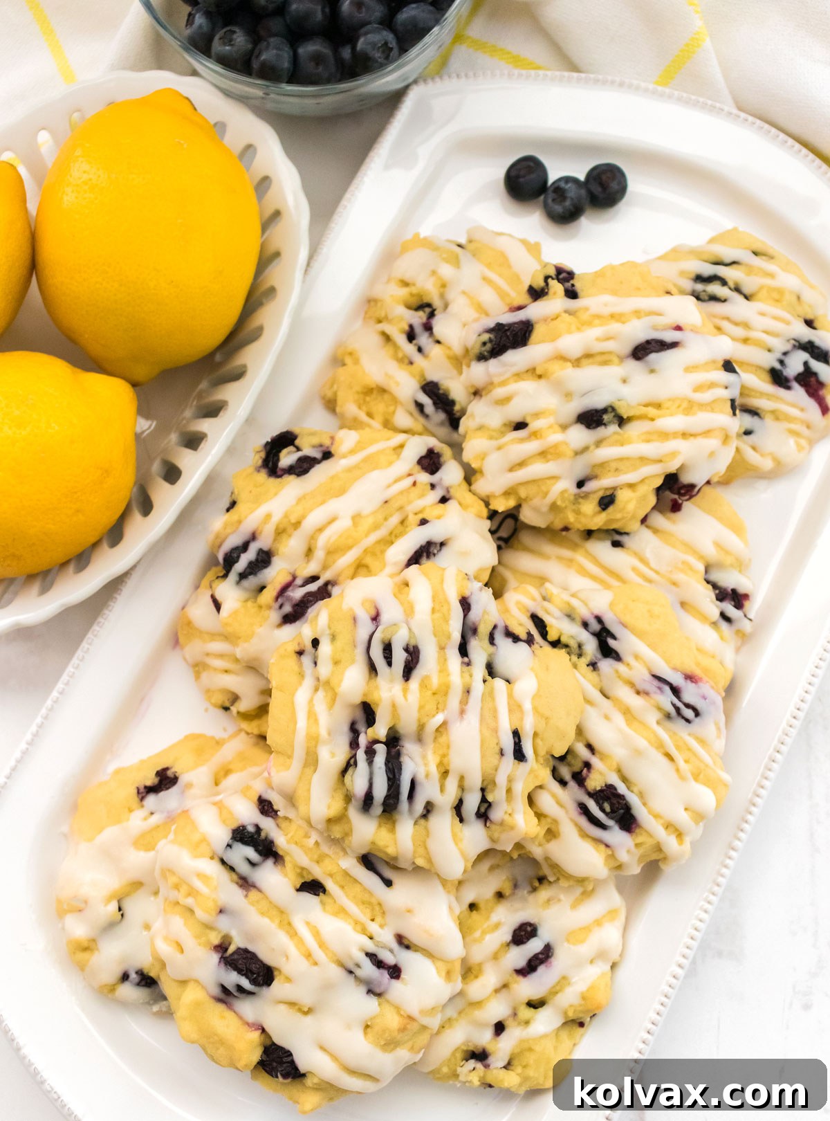 Overhead shot of a white serving platter filled with glistening Lemon Blueberry Cookies sitting next to a bowl of fresh lemons and a bowl of fresh blueberries, ready to be enjoyed.