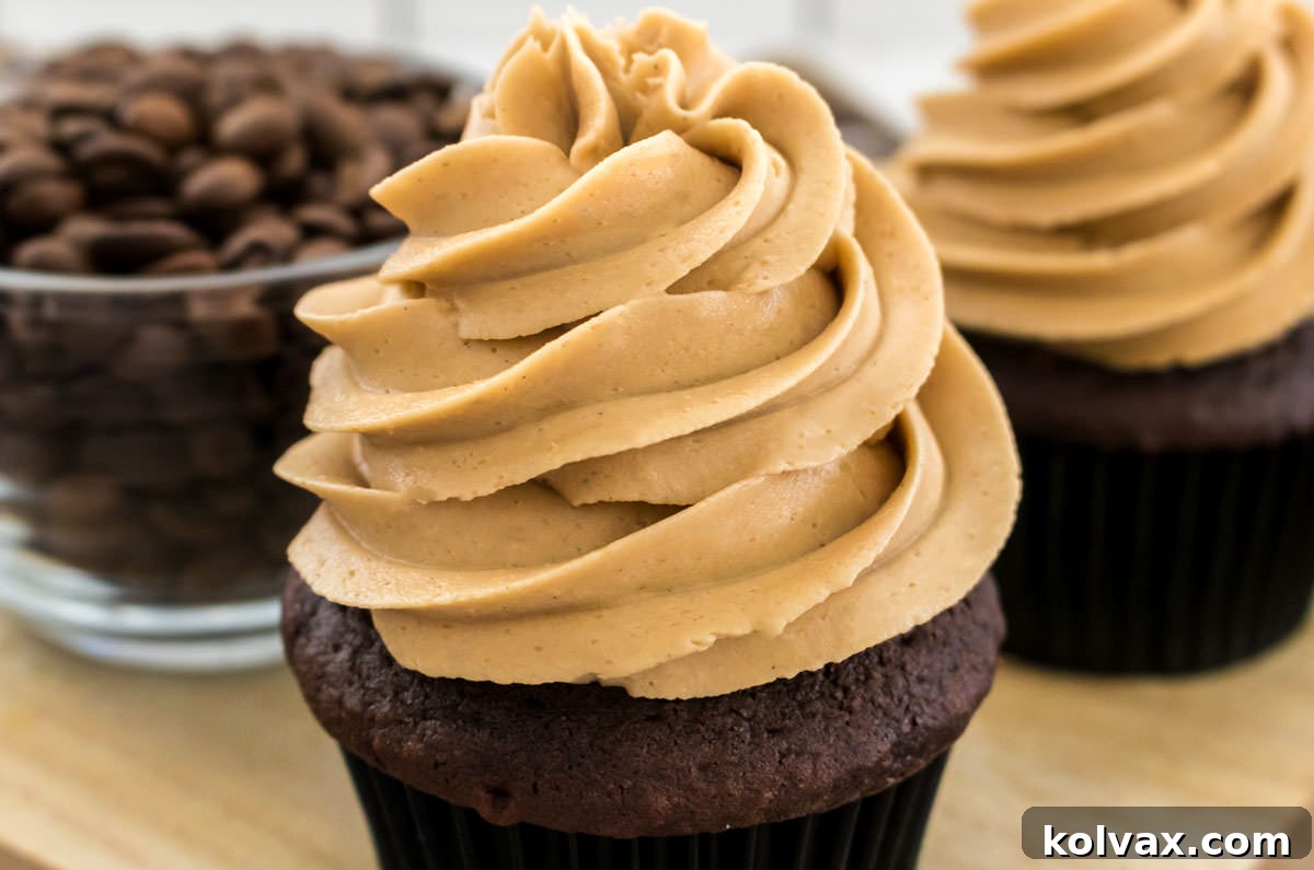 Velvet Coffee Buttercream 2 Two chocolate cupcakes generously frosted with homemade coffee buttercream, adorned with a few coffee beans, sitting on a wooden cutting board with a glass bowl of coffee beans in the background.