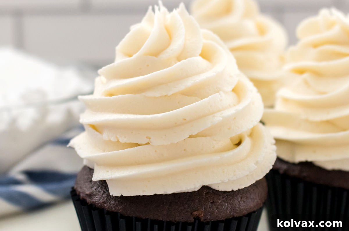 Closeup on three cupcakes topped with The Best Whipped Vanilla Frosting sitting on white table next to a glass bowl filled with powdered sugar.