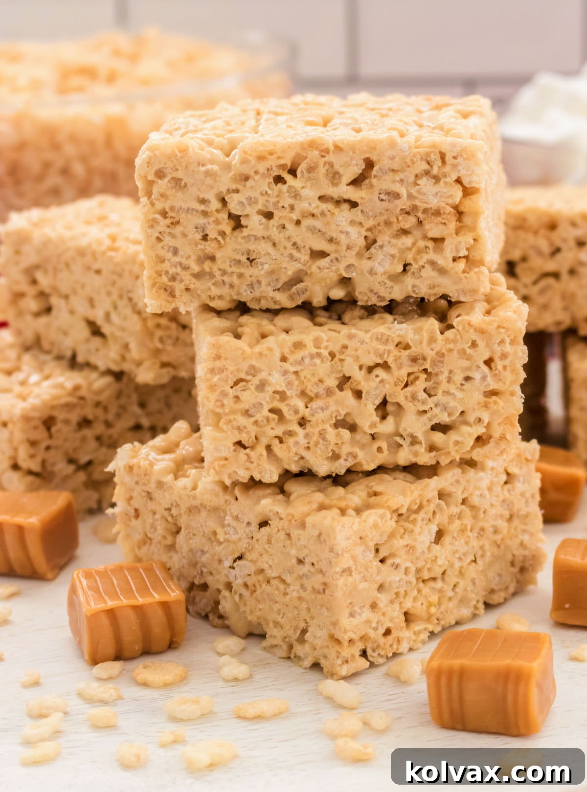 Closeup on a stack of three Caramel Rice Krispie Treats sitting on a white table surrounded by caramel candies and Rice Krispie Cereal.