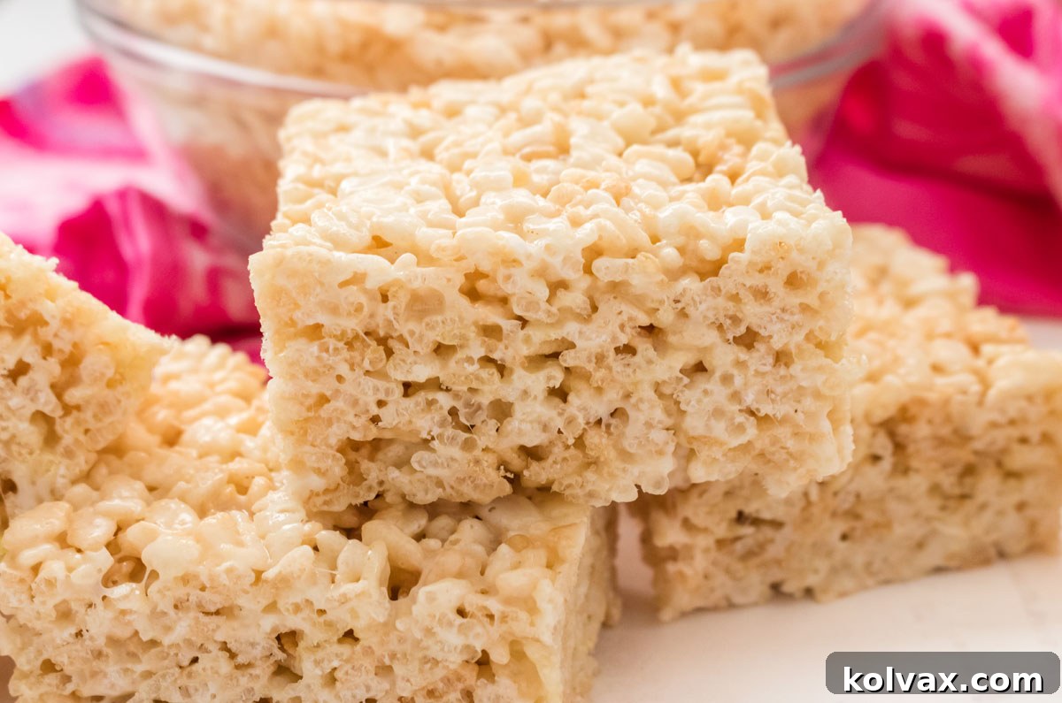Three Rice Krispie Treats sitting on a white table in front of a glass bowl filled with Rice Krispie Cereal.