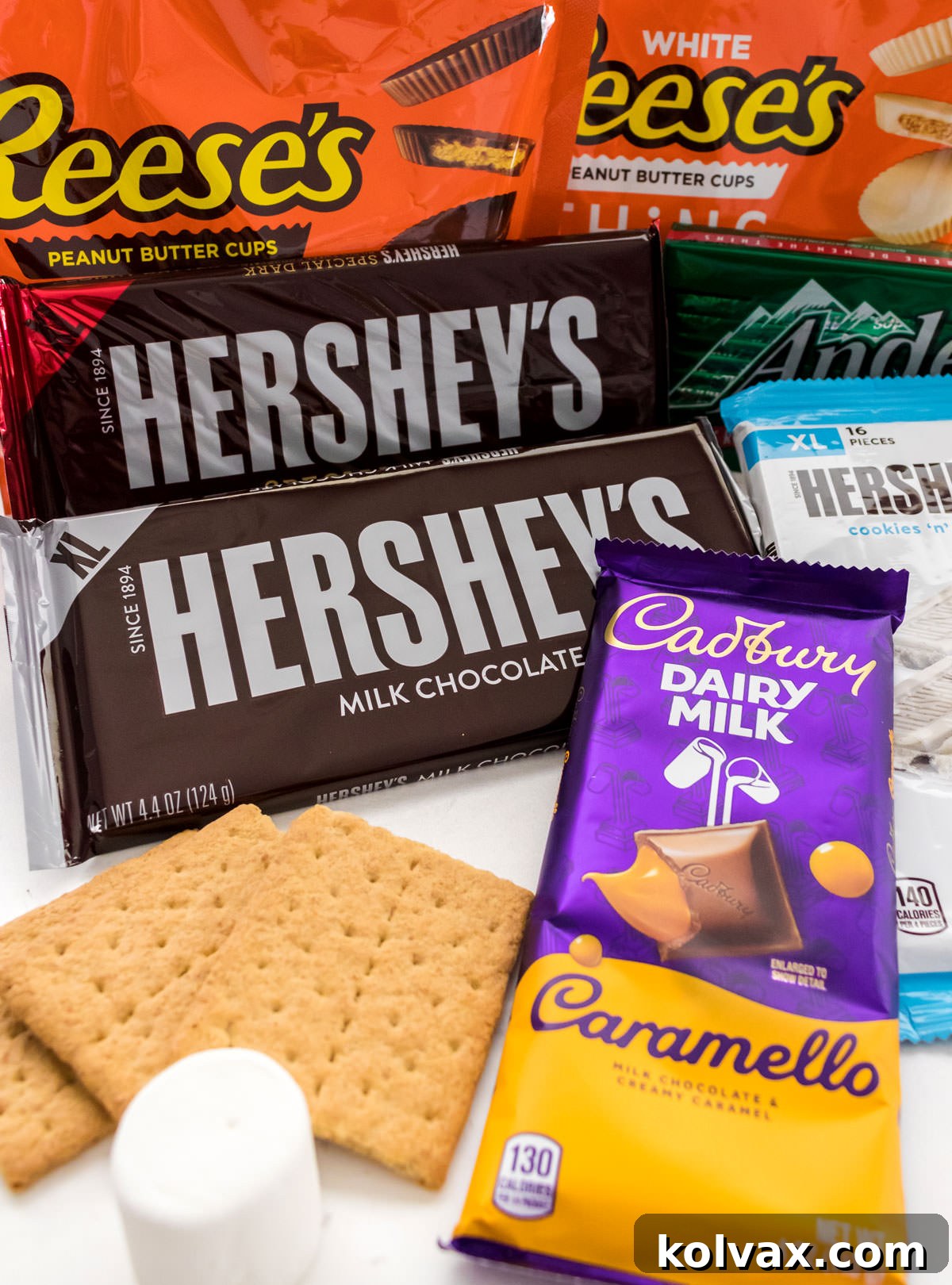 An assortment of chocolate candy bars, including Hershey's Milk Chocolate, Reese's Peanut Butter Cups, and Andes Mints, laid out with graham crackers and a marshmallow, emphasizing choice for S'mores.