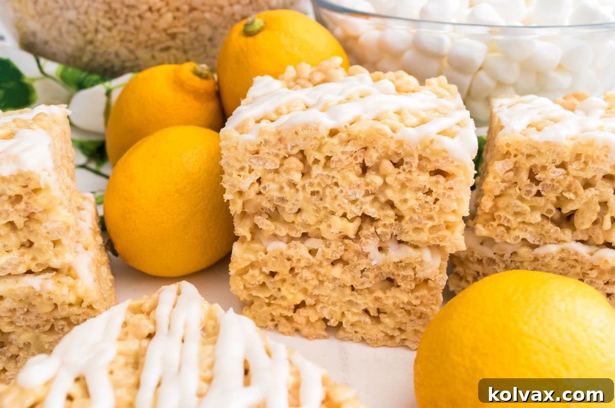 Stacks of two bright yellow Lemon Rice Krispie Treats on a white table, surrounded by fresh lemons, mini marshmallows, and rice krispie cereal. The image highlights their vibrant color and inviting texture.