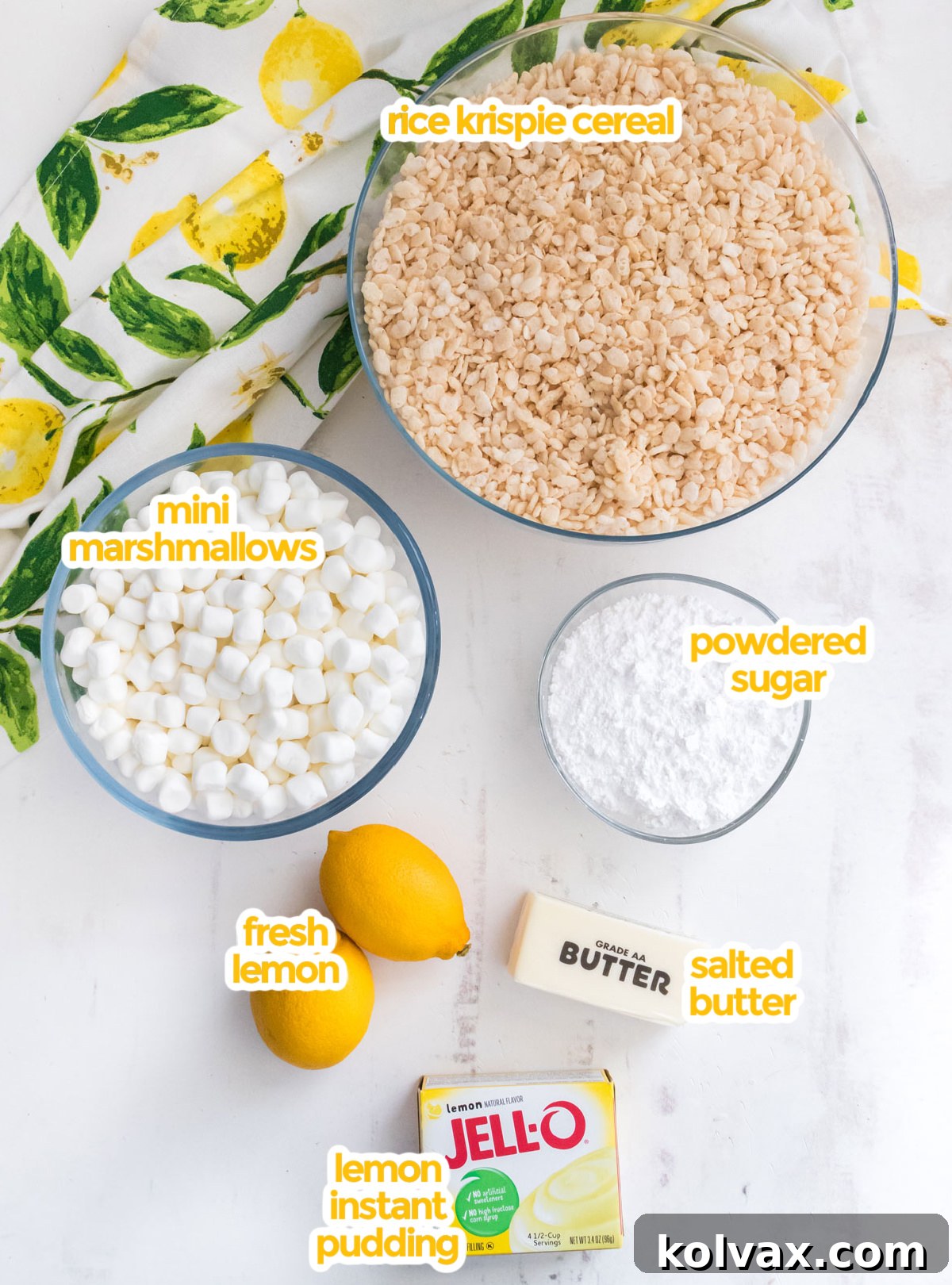 Overhead shot showcasing all the fresh and packaged ingredients laid out for making Lemon Rice Krispie Treats, including vibrant fresh lemons, blocks of butter, bags of mini marshmallows, a box of Rice Krispie cereal, powdered sugar, and a package of instant lemon pudding mix.