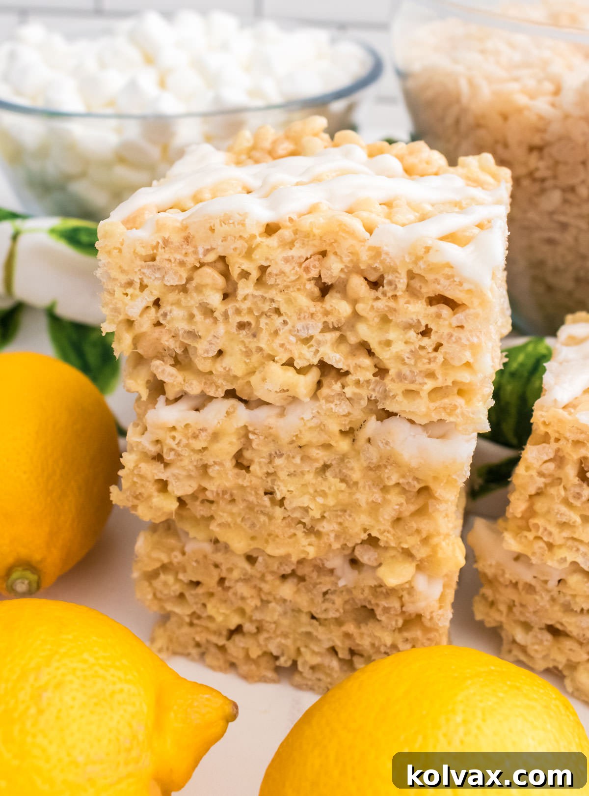 Vertical side shot of three Lemon Rice Krispie Treats stacked neatly on a white table, surrounded by bowls of mini marshmallows, rice krispie cereal, and fresh lemons, emphasizing their height, layers, and appealing texture.