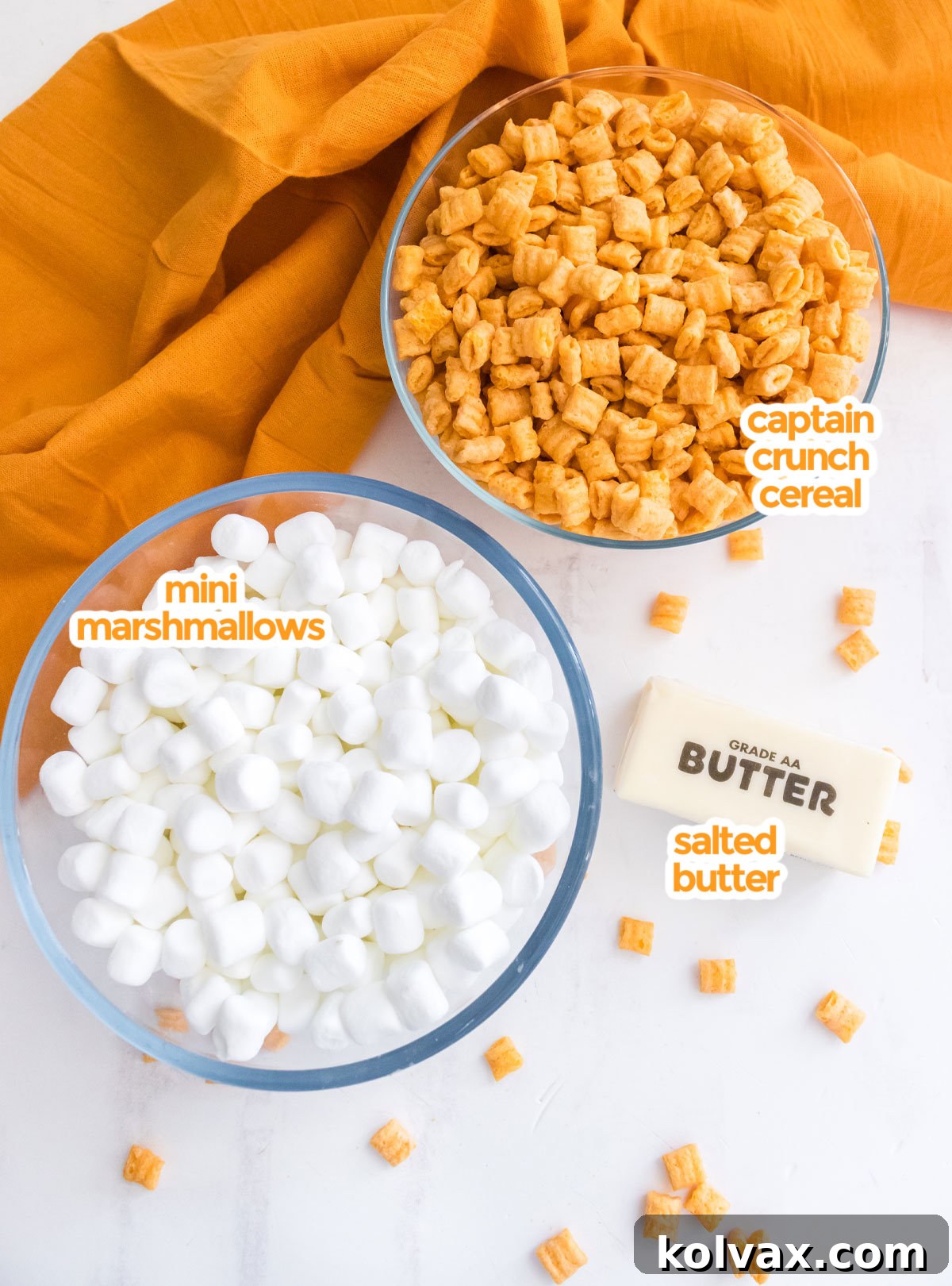 All the necessary ingredients laid out on a white surface, featuring a box of Cap'n Crunch Cereal, a bag of mini marshmallows, and a stick of butter, ready for making delicious treats.