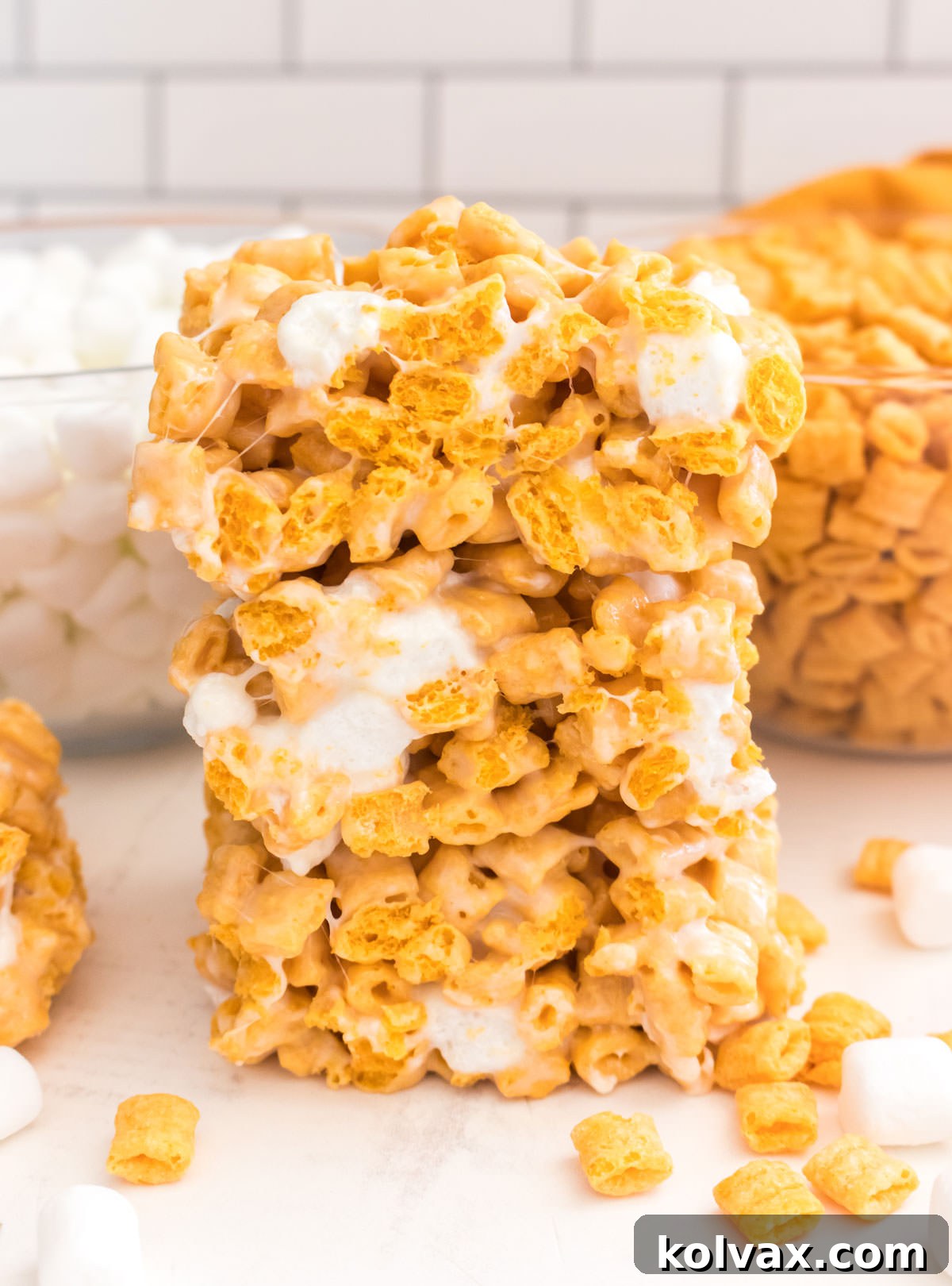 A vertical image showcasing a tempting stack of three Cap'n Crunch Marshmallow Treats on a white table, with bowls of the signature cereal and mini marshmallows blurred in the background, highlighting the irresistible dessert.