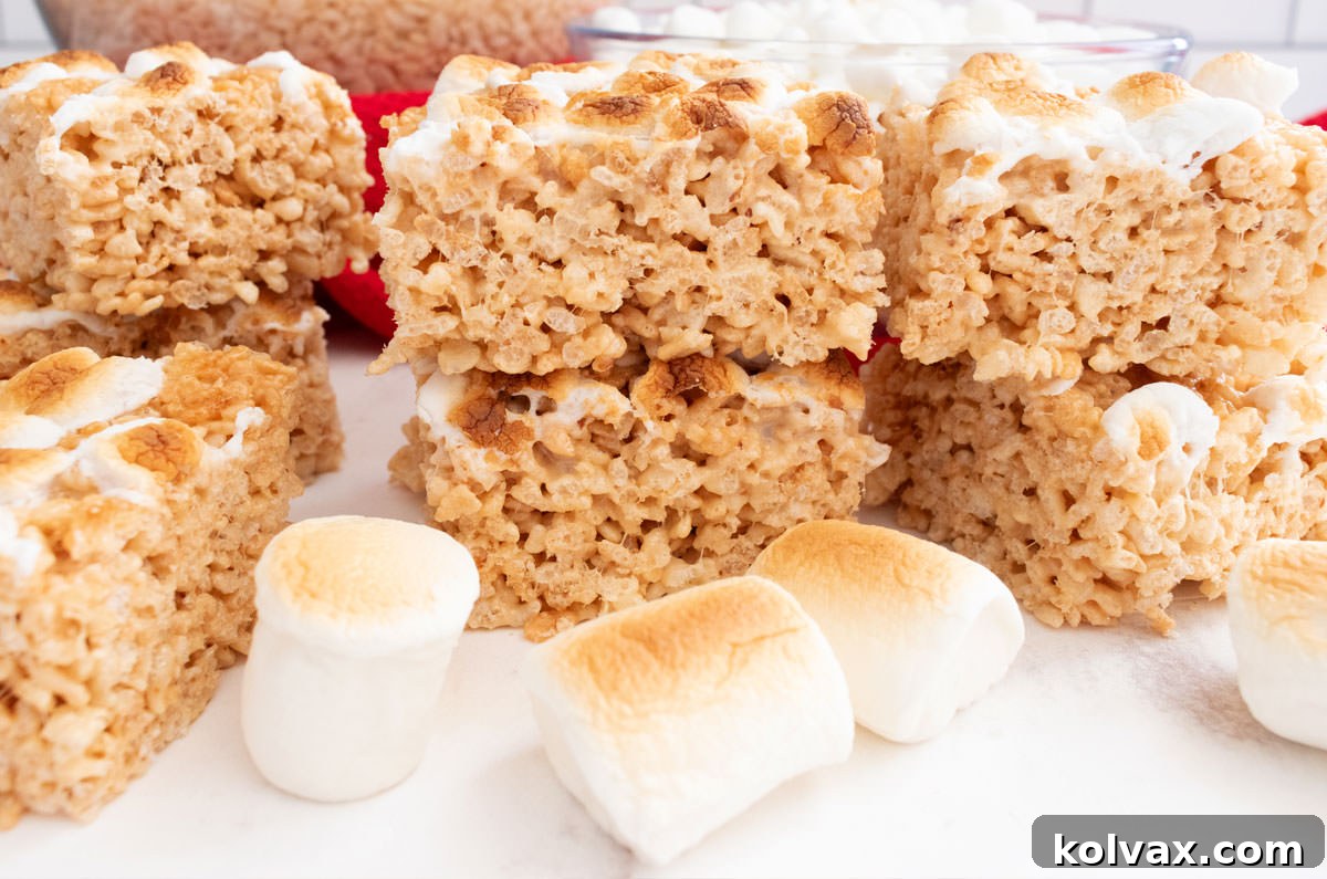 A batch of golden-brown Toasted Marshmallow Rice Krispie Treats arranged on a white table, surrounded by fluffy roasted marshmallows and crisp rice cereal, ready to be enjoyed.