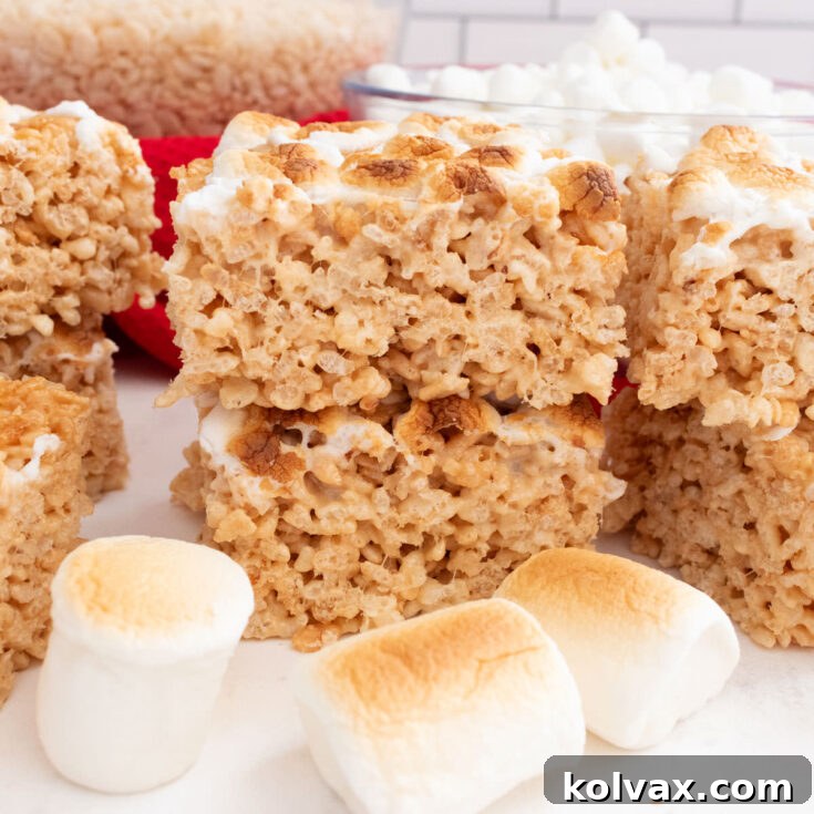 A batch of Toasted Marshmallow Rice Krispie Treats sitting on a white table surrounded by roasted marshmallows, featuring a golden-brown top layer.