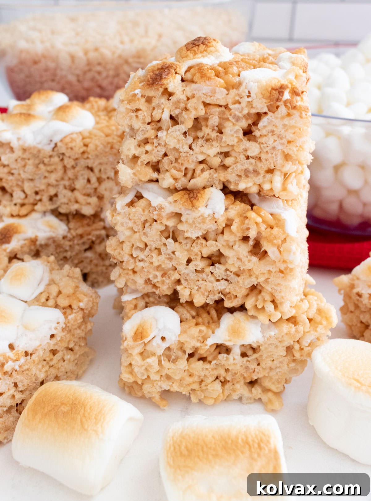 A striking vertical image of a stack of golden-brown Toasted Marshmallow Rice Krispie Treats resting on a white table, positioned in front of elegant glass bowls filled with crisp cereal and fluffy mini marshmallows.