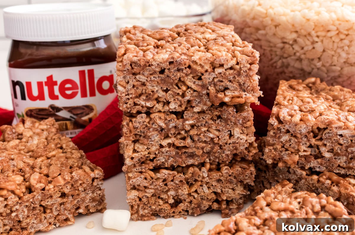 A batch of Nutella Rice Krispie Treats sitting on a white table next to a jar of Nutella Spread and a bowl of Rice Krispies cereal.