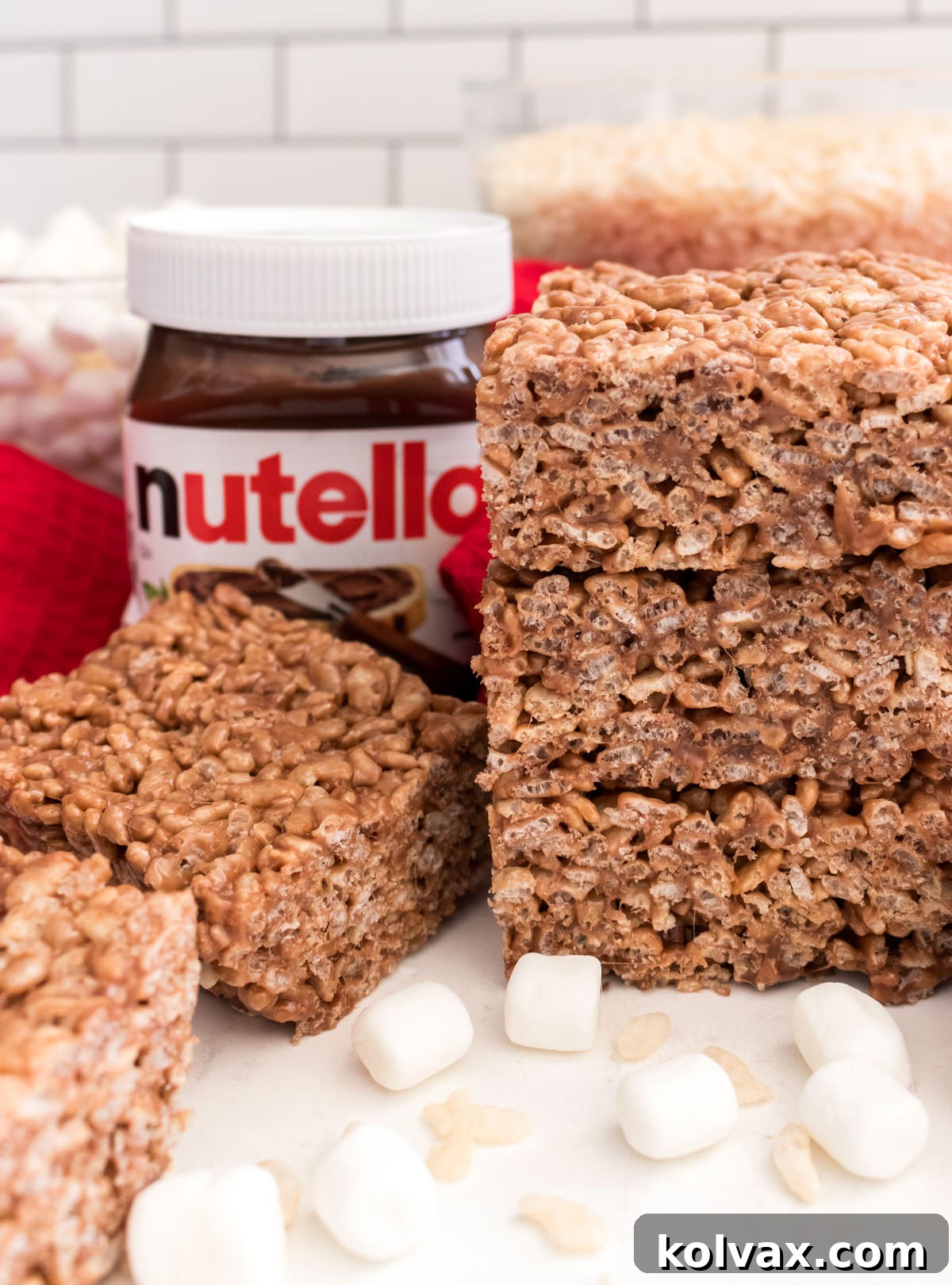 A stack of three Nutella Rice Krispie Treats sitting on a white table next to a jar of Nutella and bowl of Mini Marshmallows.