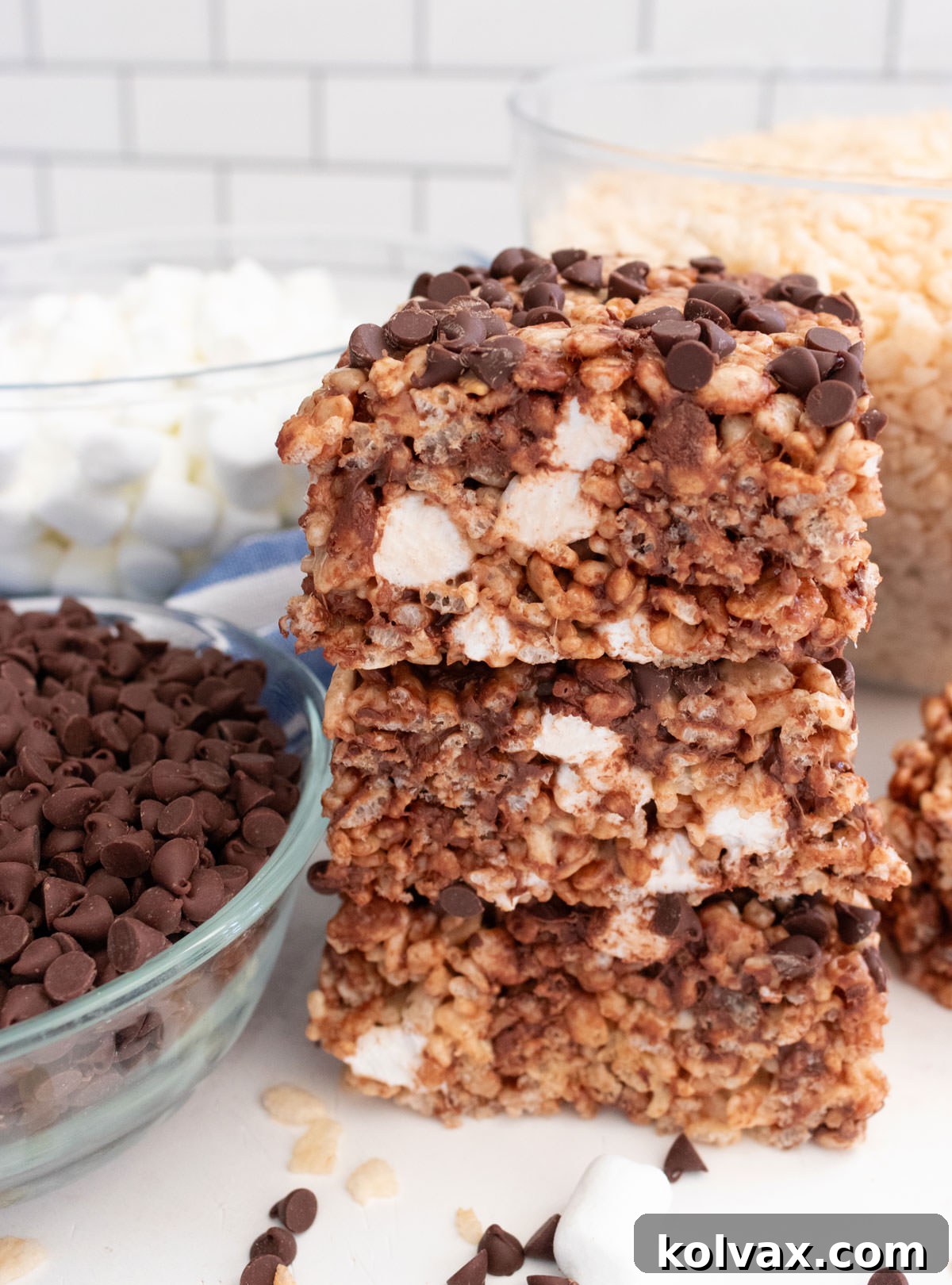 A close-up shot of a perfectly stacked trio of Zebra Rice Krispie Treats, displaying their texture and chocolate chip distribution, resting on a white surface next to a small bowl of glistening mini chocolate chips.