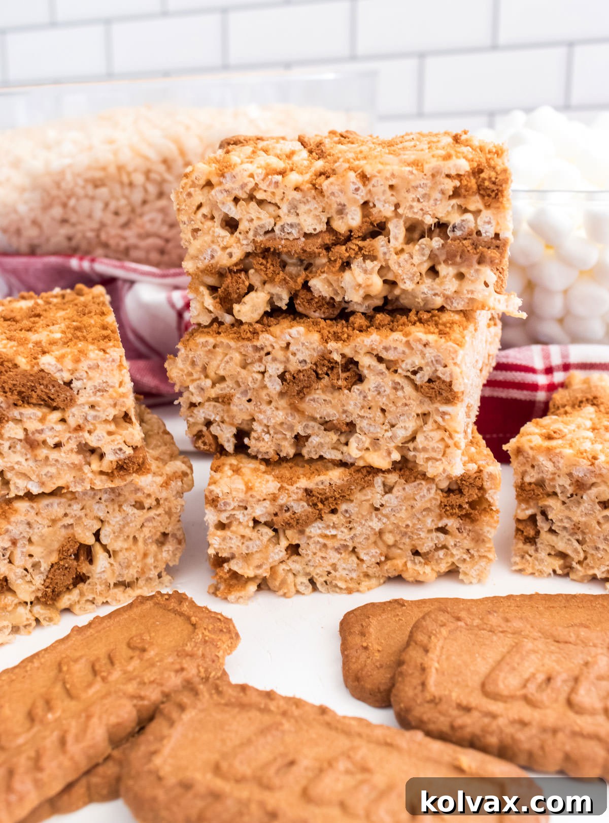 A close-up shot of three stacked Cookie Butter Rice Krispie Treats on a white table, with bowls of cereal and marshmallows blurred in the background, highlighting the delicious texture.