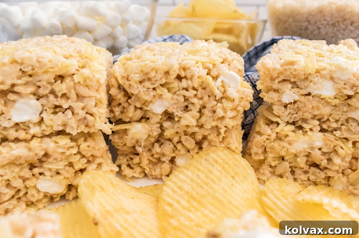 Six Potato Chip Rice Krispie Treats sitting on a white table surrounded by glass bowls filled with marshmallows and potato chips. These delectable treats combine the classic sweetness of marshmallows with the savory crunch of potato chips for a unique and satisfying dessert.