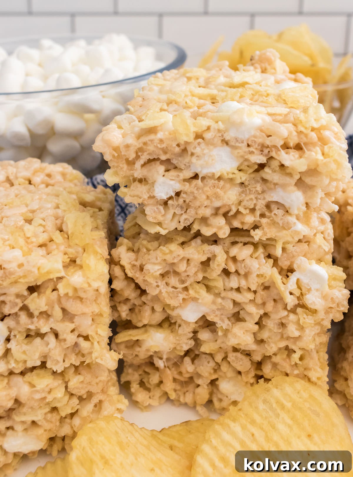 A close-up of two neat stacks of Potato Chip Rice Krispie Treats, showcasing their crunchy texture, resting on a white table in front of glass bowls brimming with mini marshmallows and golden potato chips.