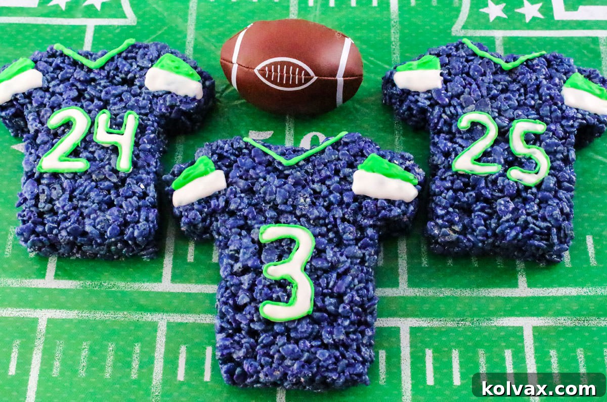 Three Seattle Seahawks Rice Krispie Treats shaped like team jerseys, neatly arranged on a vibrant green tablecloth next to a miniature toy football decoration.