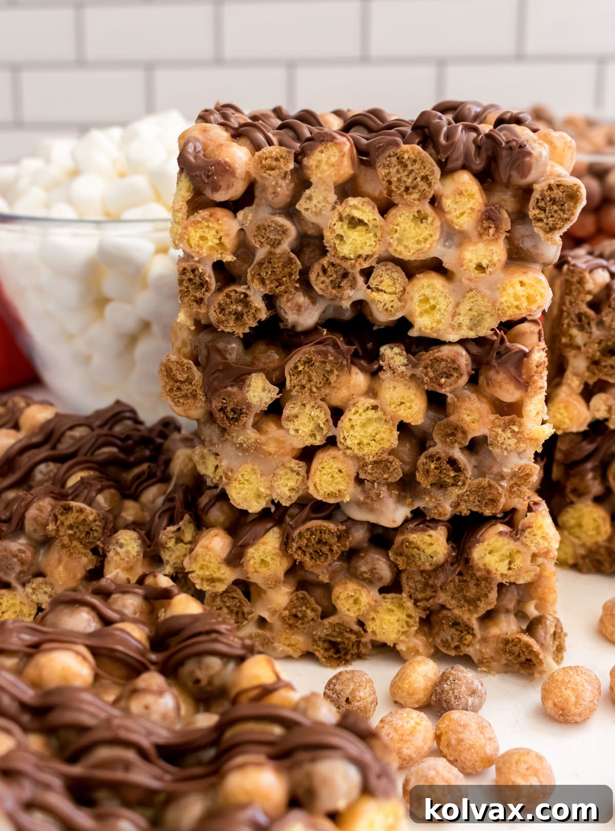 A beautiful stack of three Reese's Puffs Marshmallow Treats, showcasing their delicious layers, sitting on a white table surrounded by loose Reese's Puffs cereal and mini marshmallows, highlighting the ingredients.