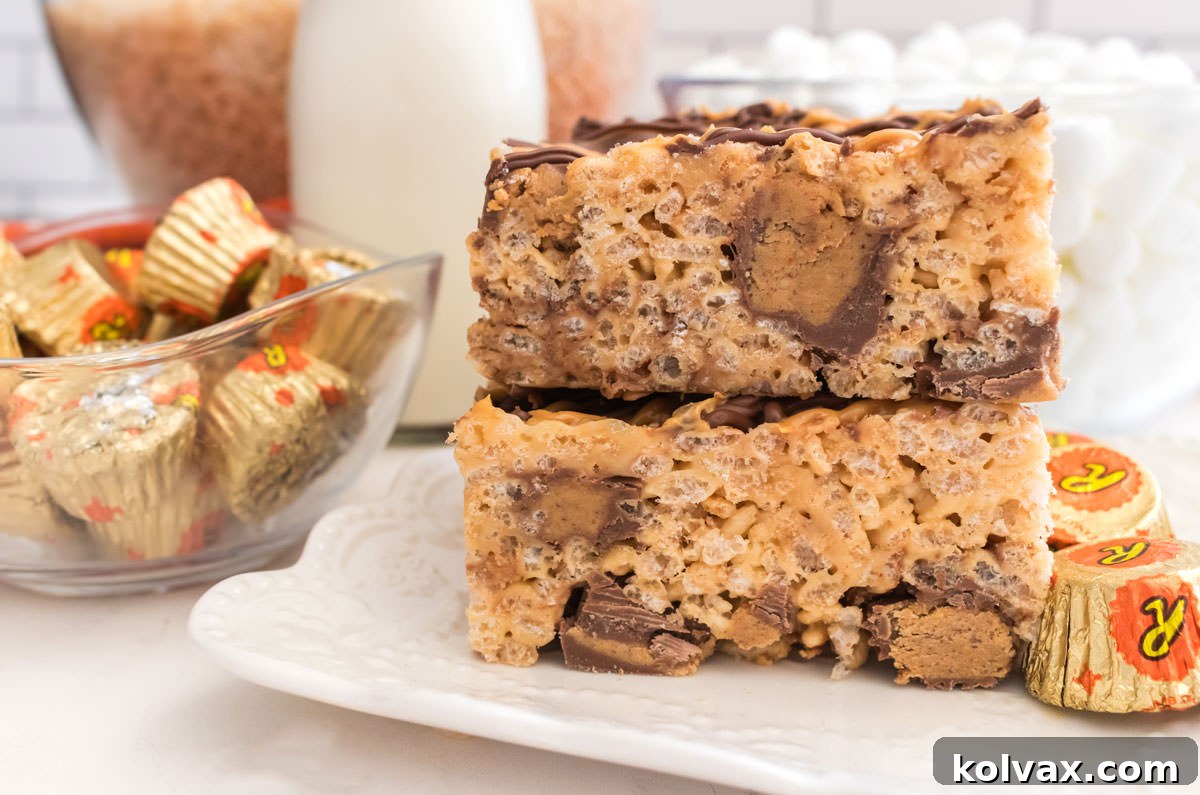 Two Peanut Butter Cups Rice Krispie Treats sitting on a white dessert plate next to a glass bowl filled with Reese's Peanut Butter Cups, showcasing their rich texture and peanut butter goodness.