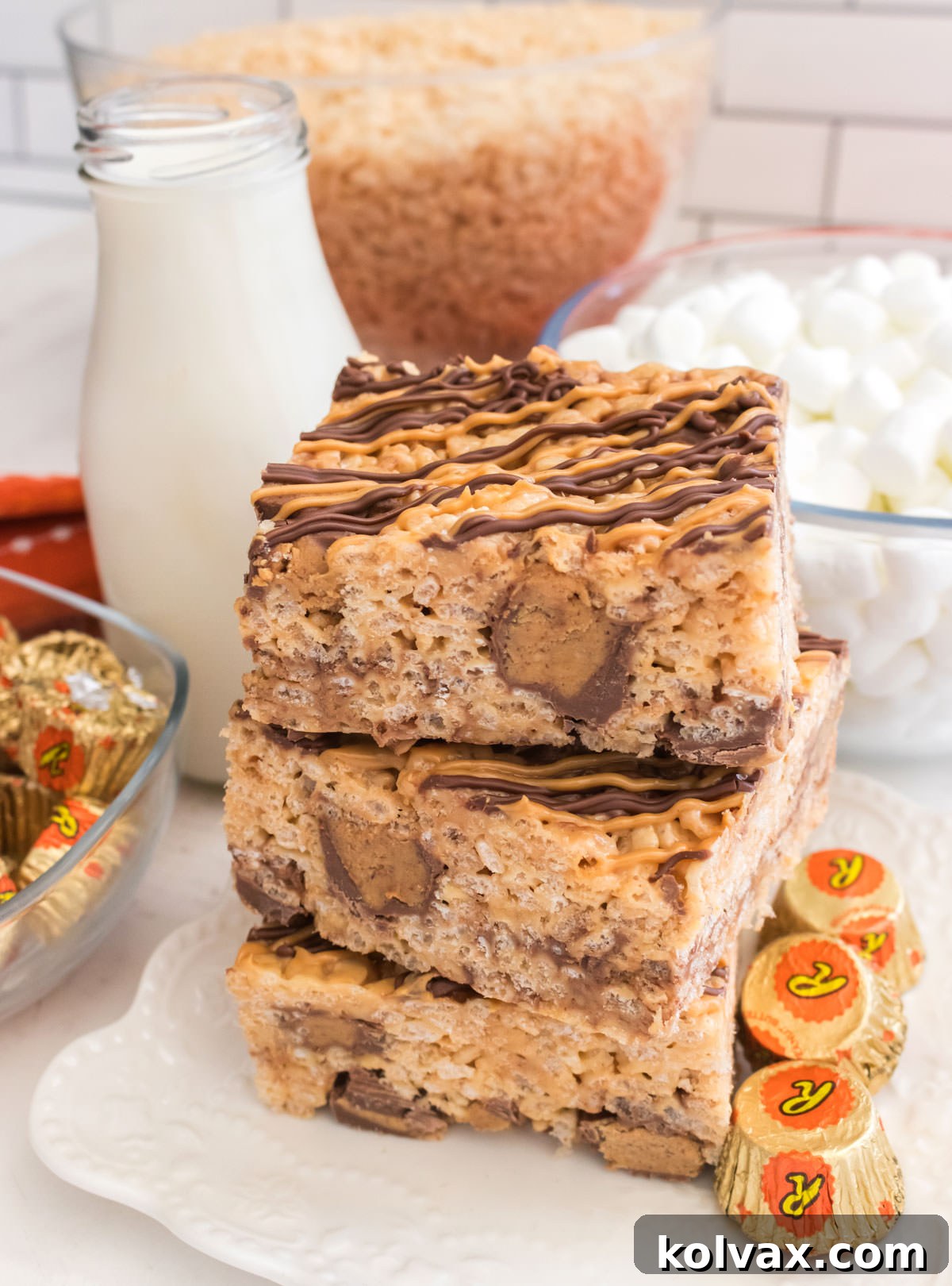A stack of three Peanut Butter Cup Rice Krispie Treats on a white dessert plate, accompanied by a glass of milk and small bowls of cereal and mini marshmallows, highlighting their perfect pairing.