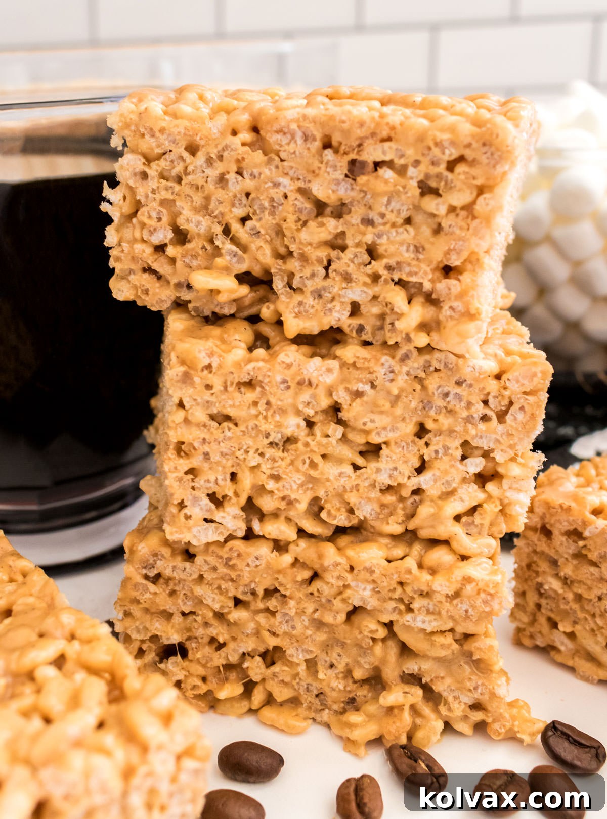 A stack of three perfectly cut Coffee Rice Krispie Treats placed next to a steaming cup of coffee on a clean white table, emphasizing their perfect pairing.