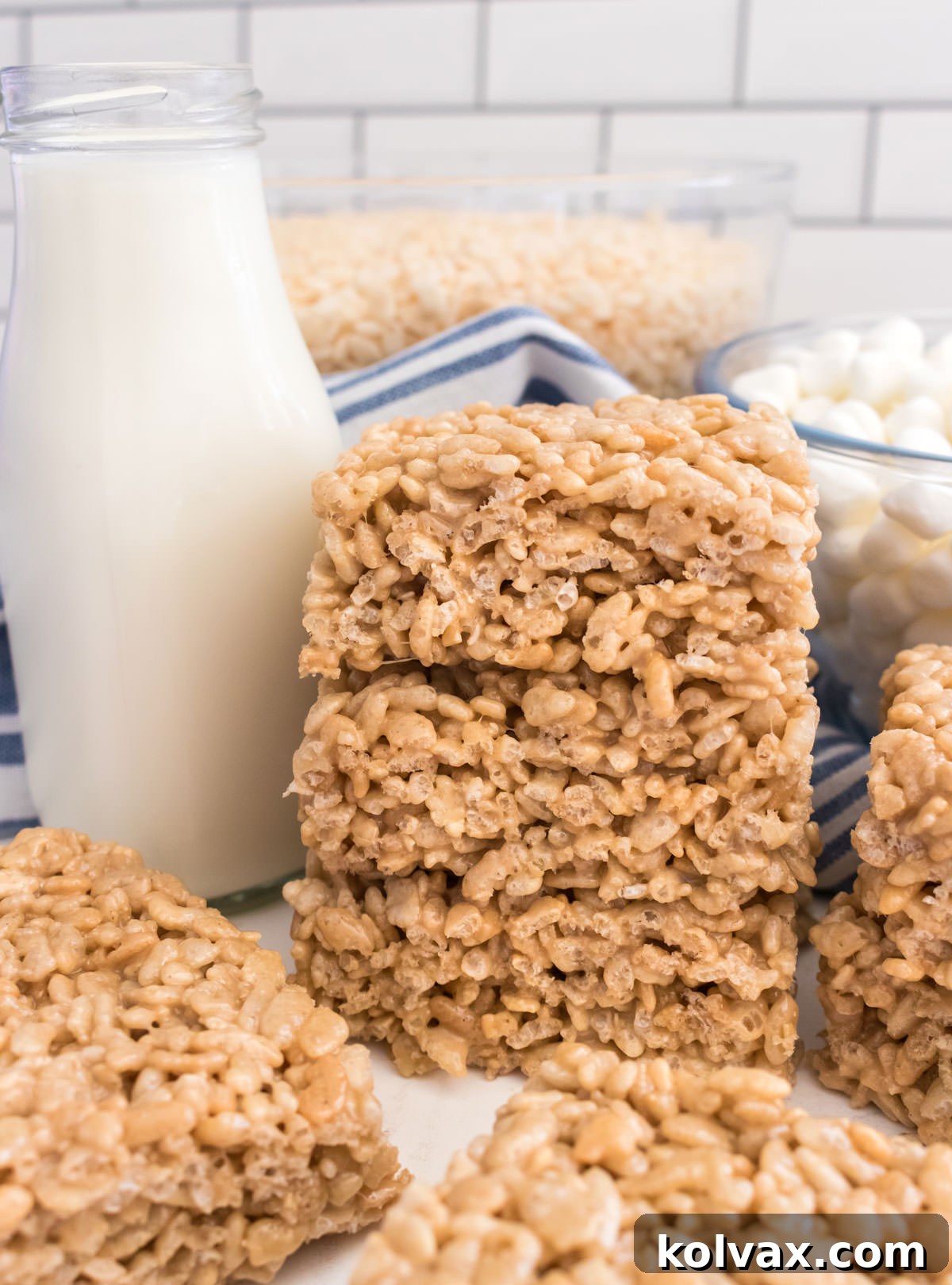 A close-up shot of a stack of three perfectly cut Maple Rice Krispie Treats, sitting invitingly on a white table next to a refreshing glass of milk, emphasizing their appealing texture.