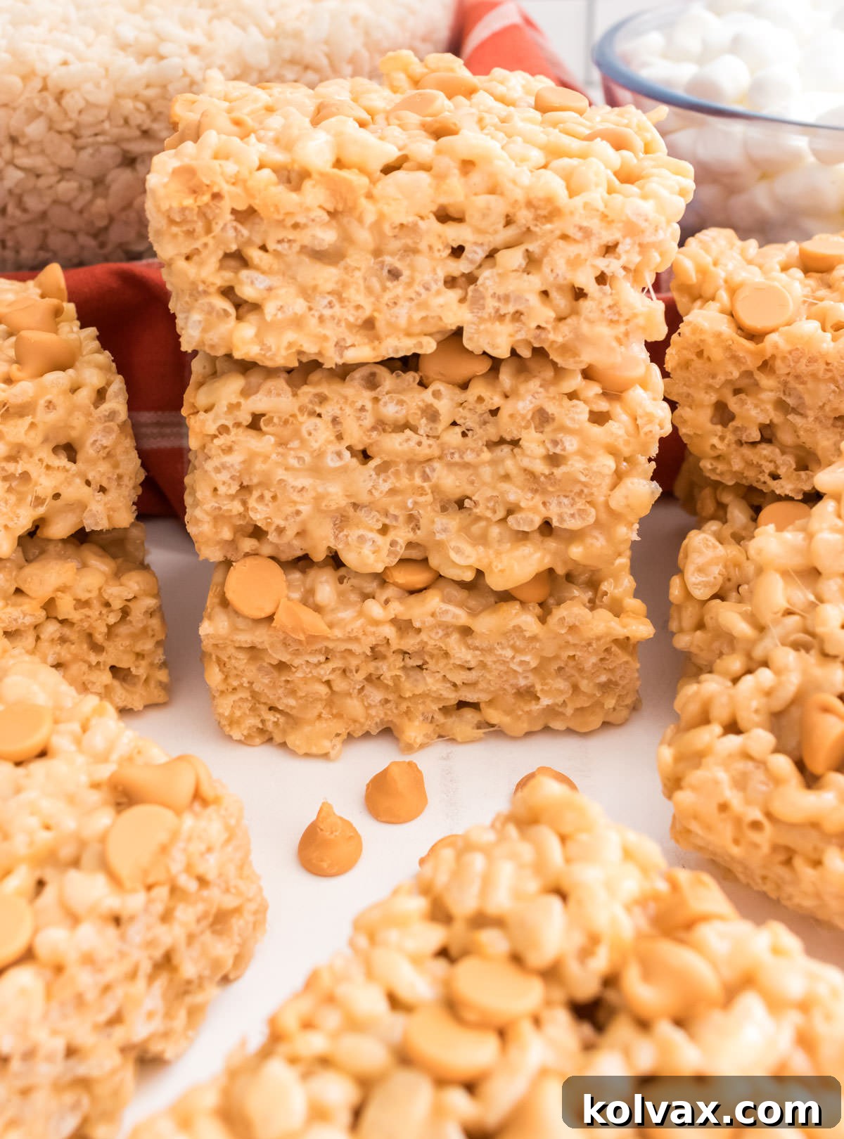 A beautiful closeup of a stack of three golden Butterscotch Rice Krispie Treats, resting on a white table. In the background, clear bowls hold Rice Krispies Cereal and Mini Marshmallows, hinting at the simple ingredients used.