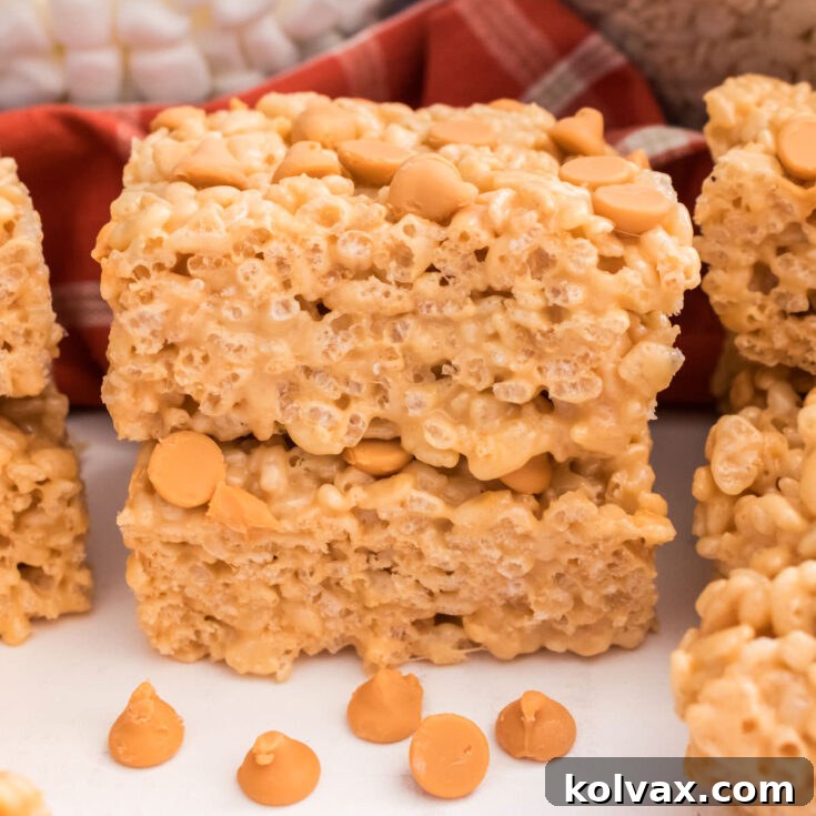 A stack of two golden Butterscotch Rice Krispie Treats sitting on a white table, surrounded by loose Butterscotch Chips and Mini Marshmallows.