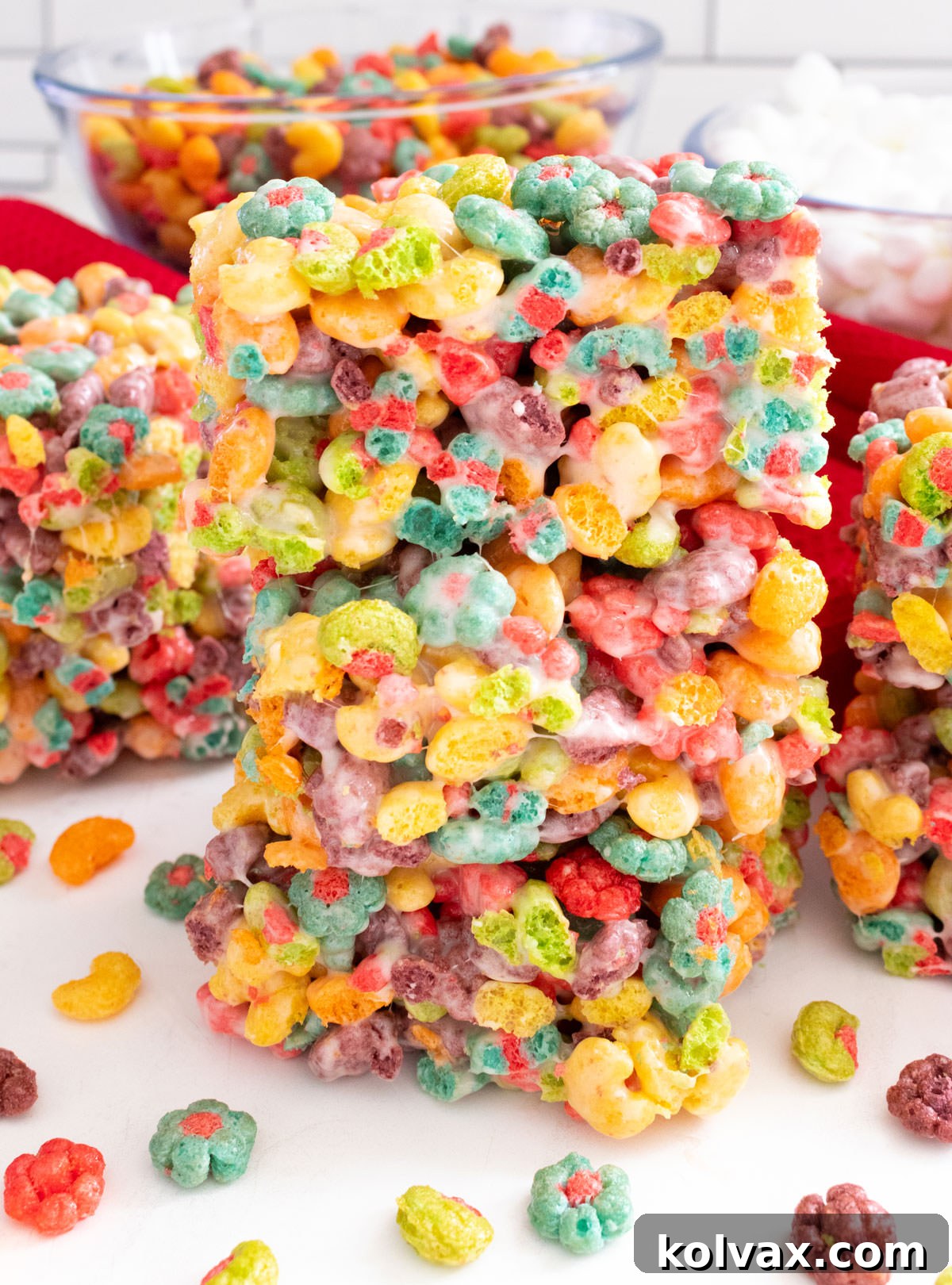 Trix Fruity Marshmallow Squares 7 A close-up of a stack of three vibrant Trix Marshmallow Treats, surrounded by loose Trix cereal on a white table.