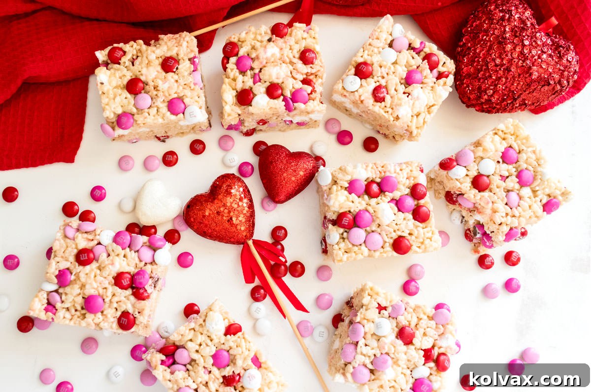 Sweetheart M&M Rice Krispie Bars 8 An inviting overhead photograph of a fresh batch of Valentine's Day M&M Rice Krispie Treats, neatly cut into squares and arranged on a white table, accompanied by a scattering of loose red, pink, and white M&M candies.