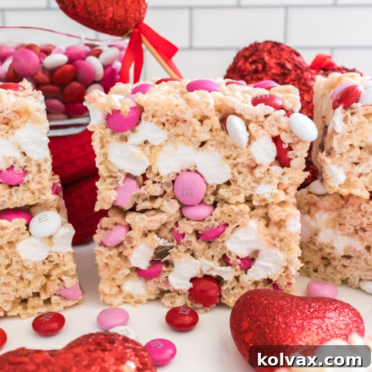 A stack of two colorful Valentine's Day M&M Rice Krispie Treats positioned next to a clear glass bowl filled with extra M&M candies, set on a pristine white table.